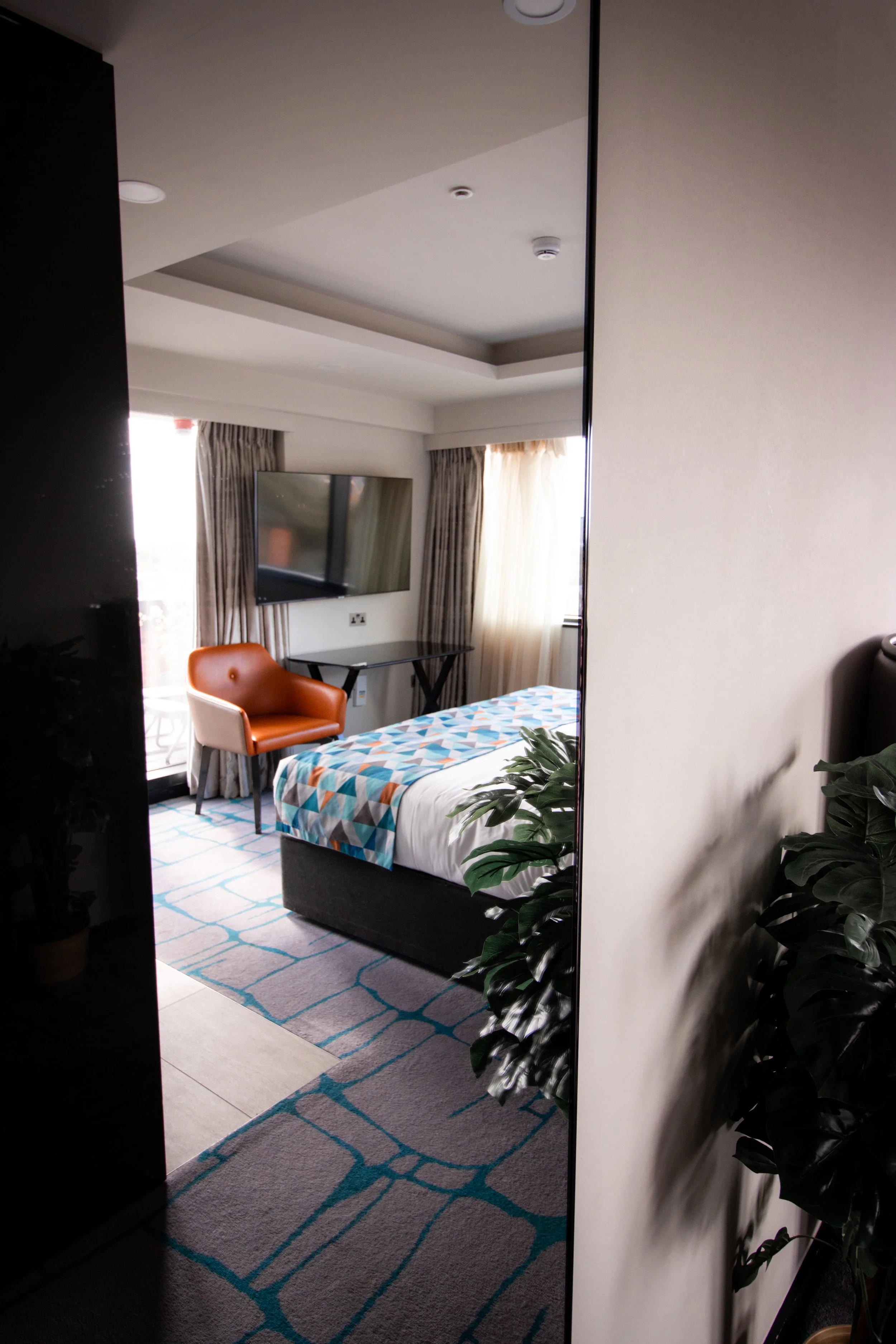 A hotel room with a bed, an orange armchair, a wall-mounted flat-screen TV, a window with curtains, a small desk, some green indoor plants, and a colorful patterned bedspread.