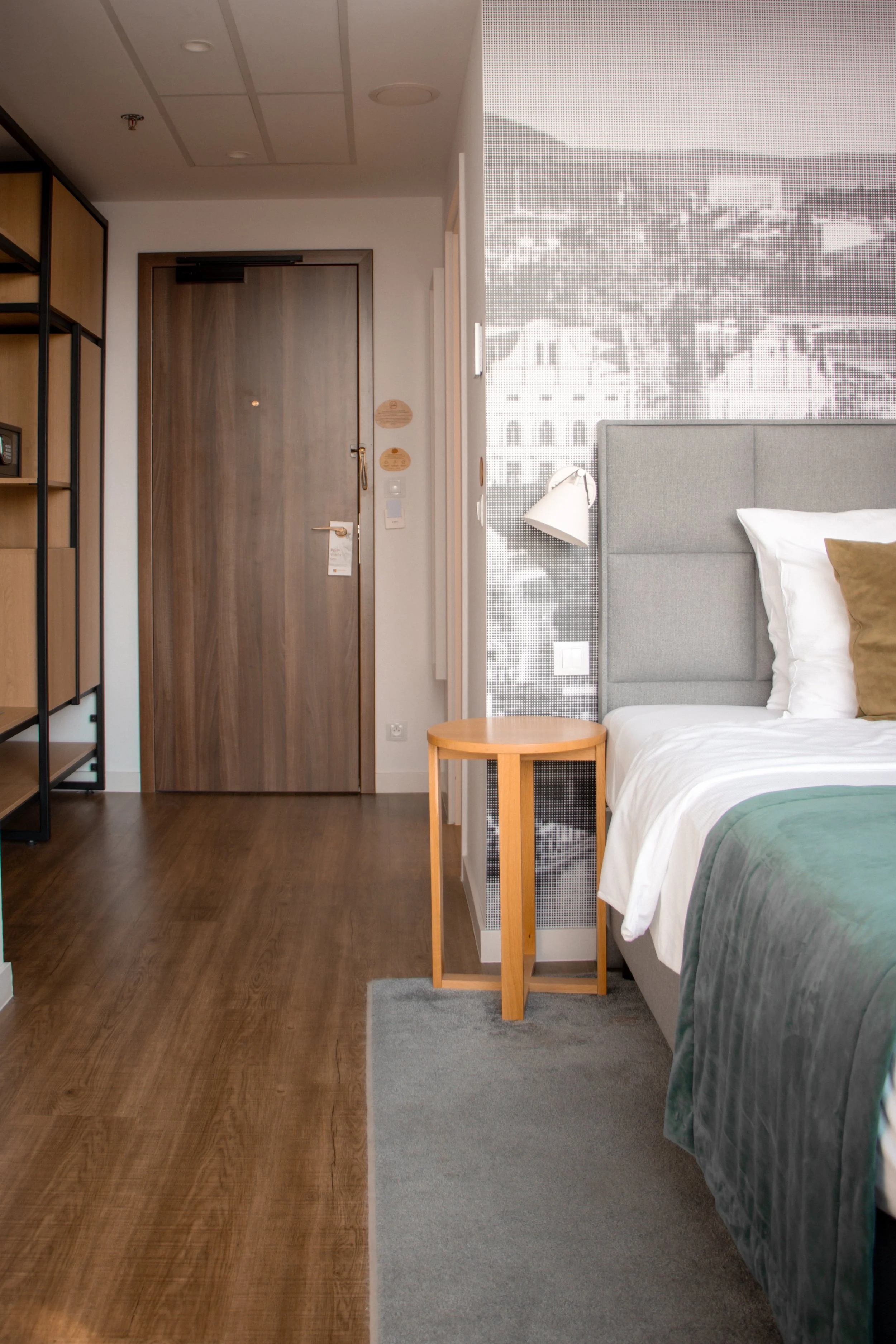 A hotel room with a gray upholstered bed, a small wooden bedside table, a white wall-mounted lamp, and a large window with a patterned blind. The room has wooden flooring and a wooden door.