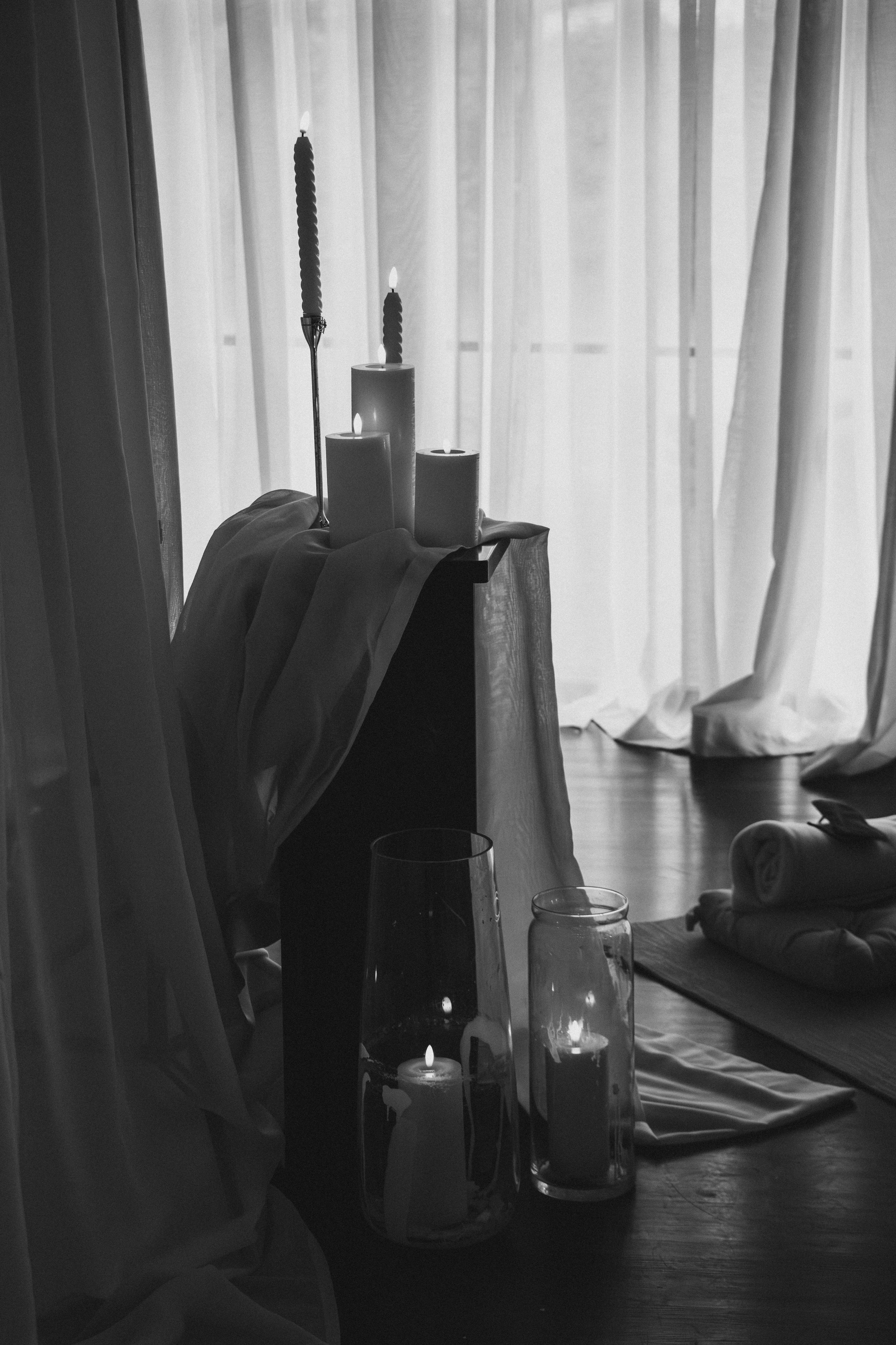 Black and white photo of lit candles on a small table near a window with sheer curtains, creating a cozy ambiance.