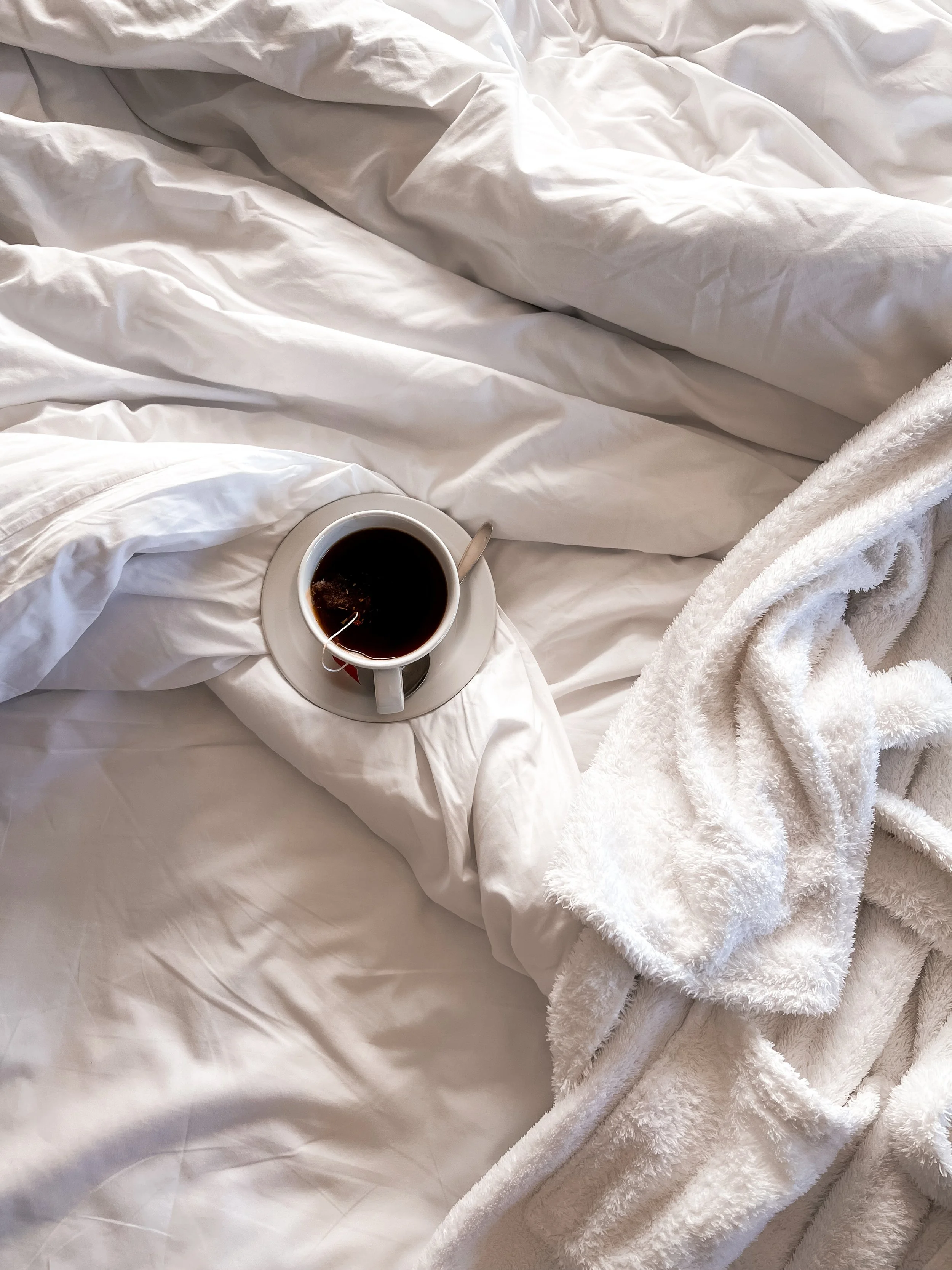 A white bed with rumpled sheets and a white fluffy blanket, with a white cup of black coffee on a saucer and a metal spoon resting on the cup, on the bed.