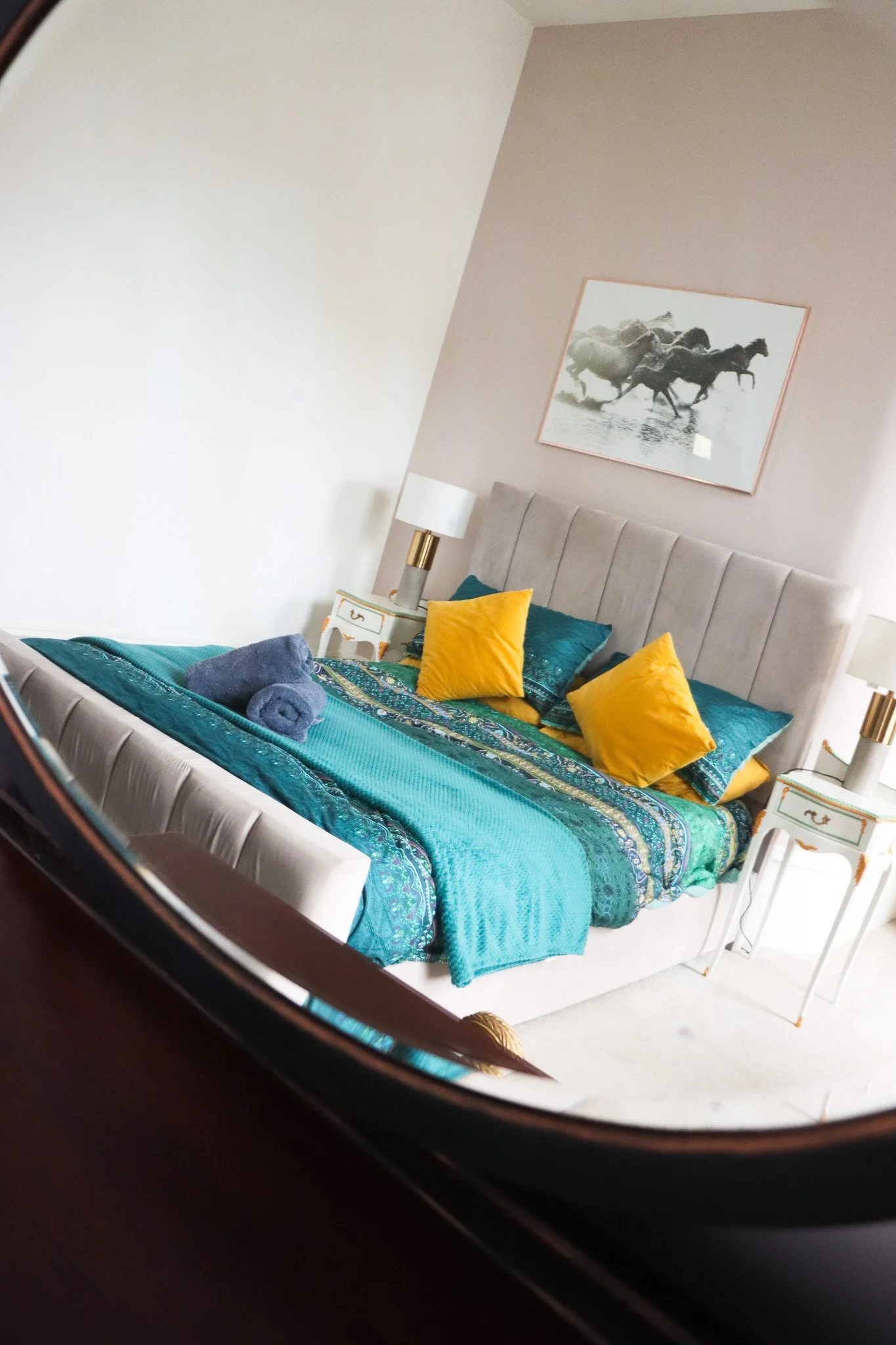 A neatly made bed with teal and yellow bedding and pillows in a modern hotel room, reflected in a mirror.