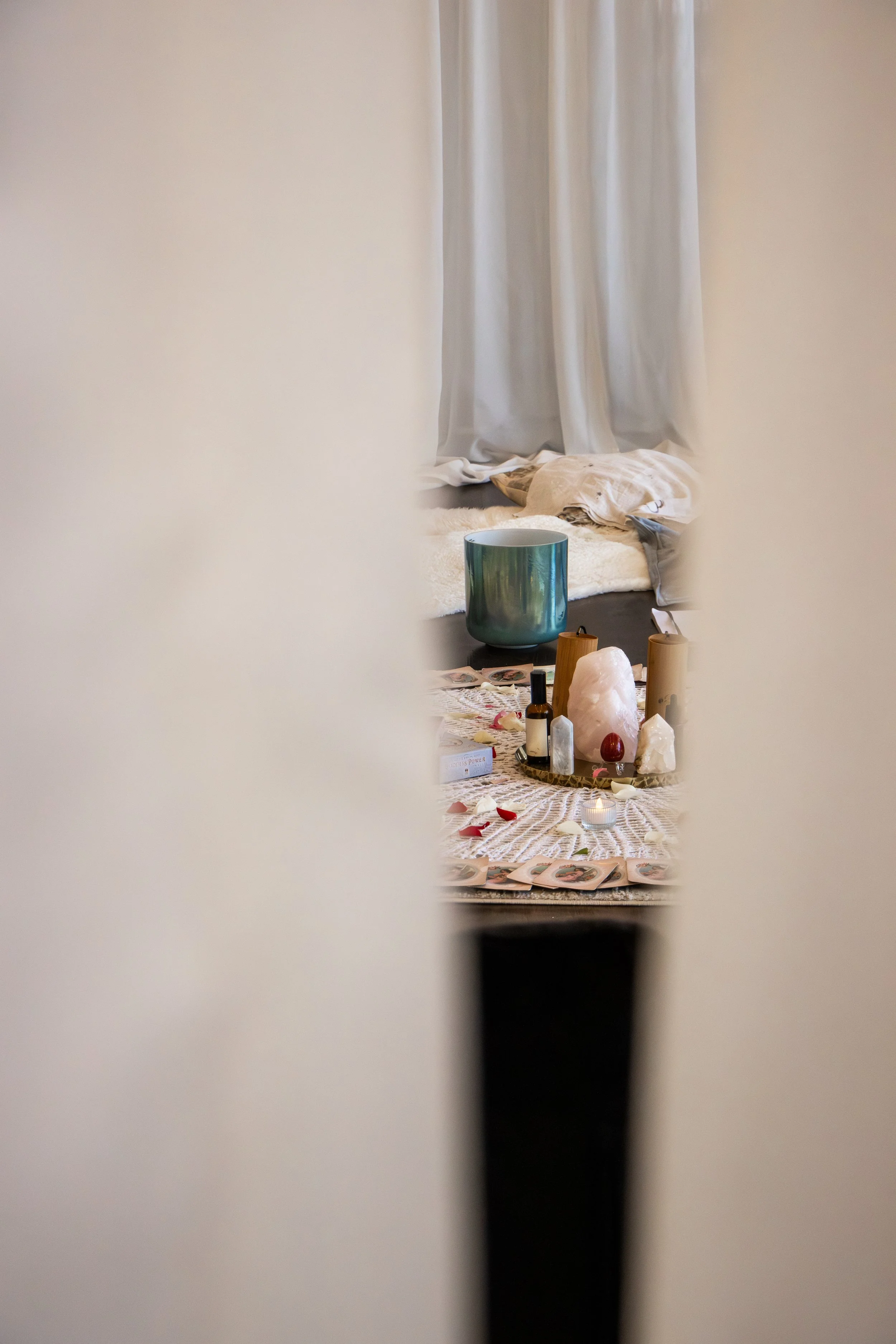 A glimpse through a narrow opening showing a workspace with candles, crystals, and tarot cards on a table, with curtains and a bed in the background.