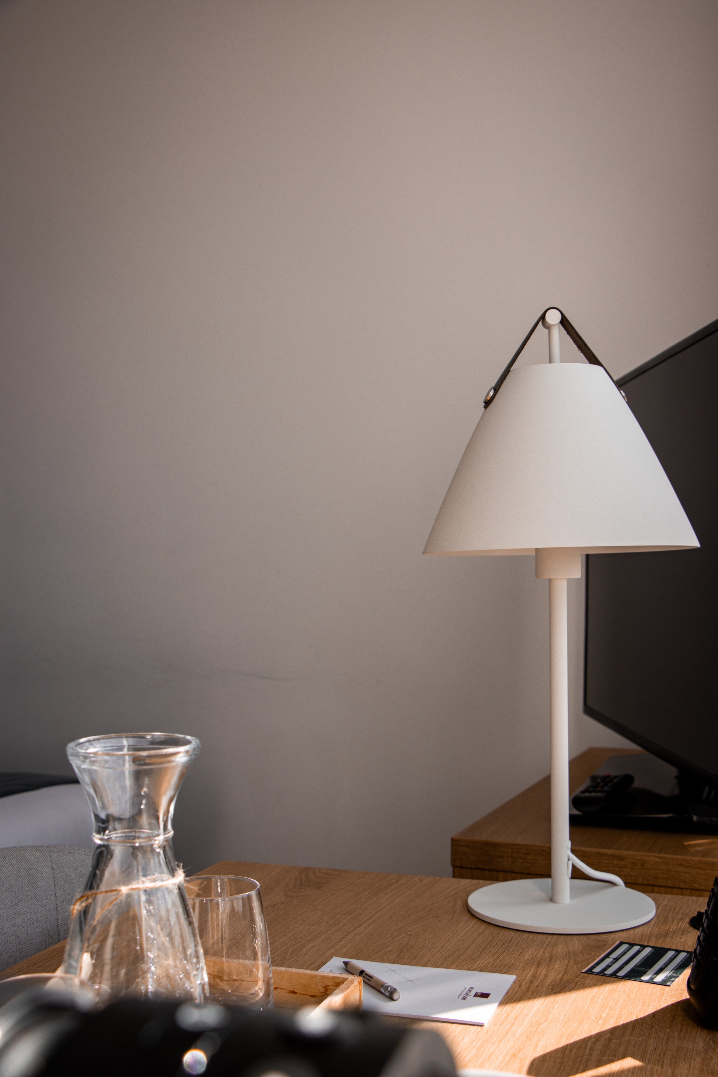 Desk with a white lamp, glass pitcher, glass, notepad, and remote control in a well-lit room.