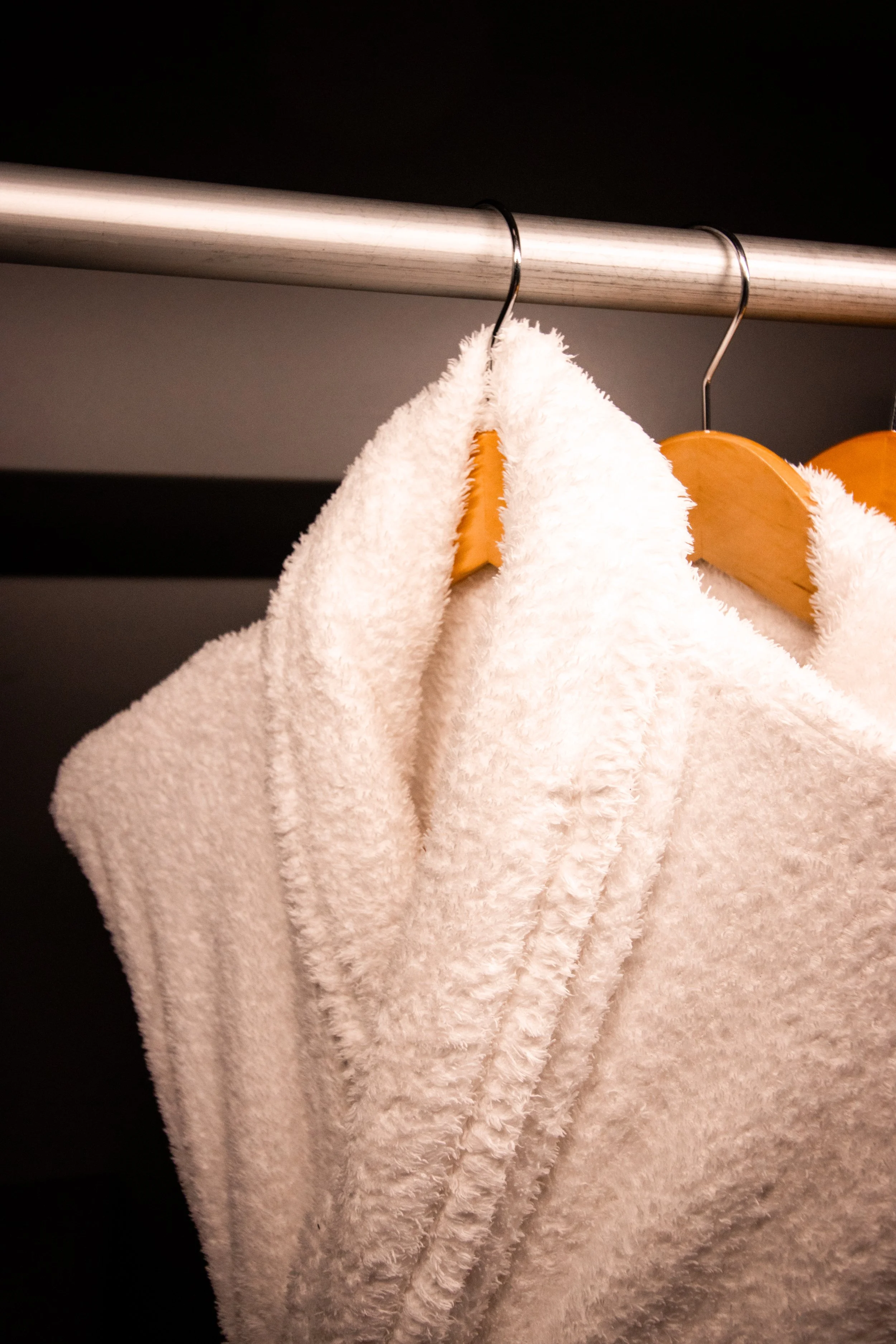 Two fluffy white bathrobes hanging on wooden hangers in a closet with a metal rod.