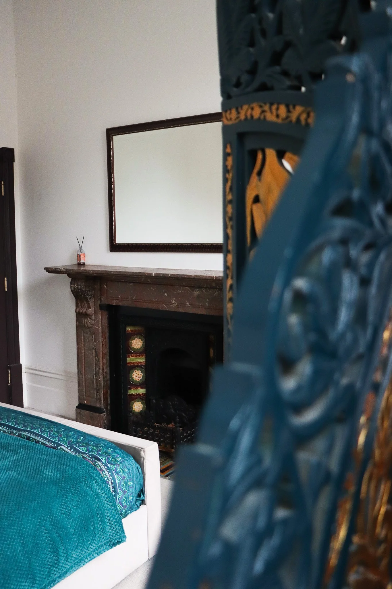 A bedroom corner with a white bed covered in a teal and blue patterned blanket, a vintage wooden fireplace mantel, a large mirror reflecting a white wall, and an ornate teal decorative panel in the foreground.