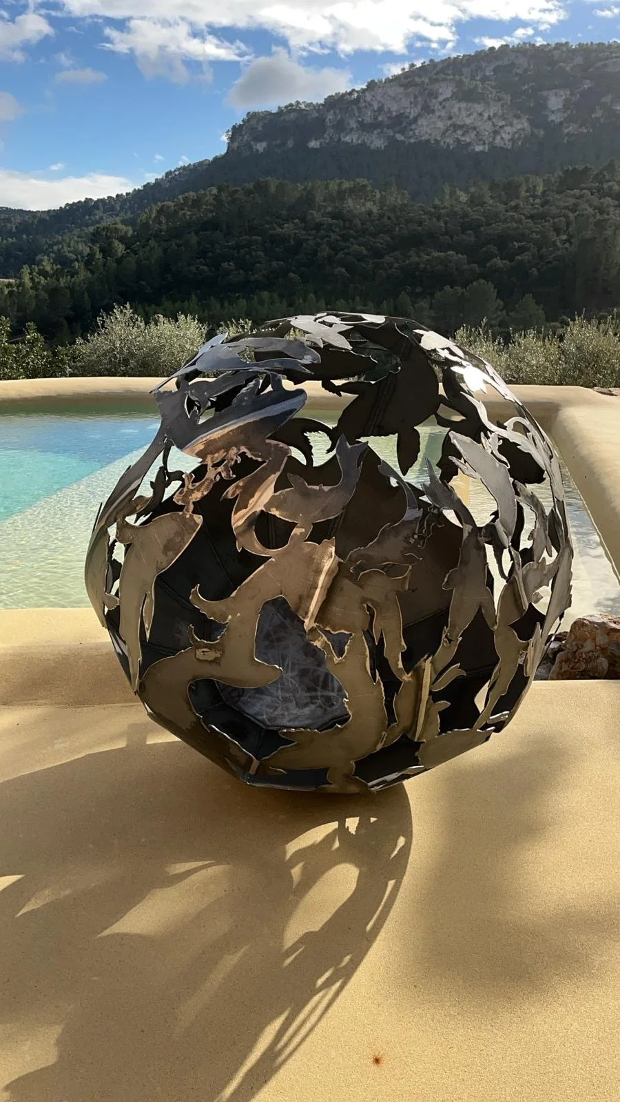 A spherical metal sculpture with intricate cut-out patterns sitting on a patio table outdoors. In the background, there is a swimming pool, green hillside, mountains, and a partly cloudy sky.