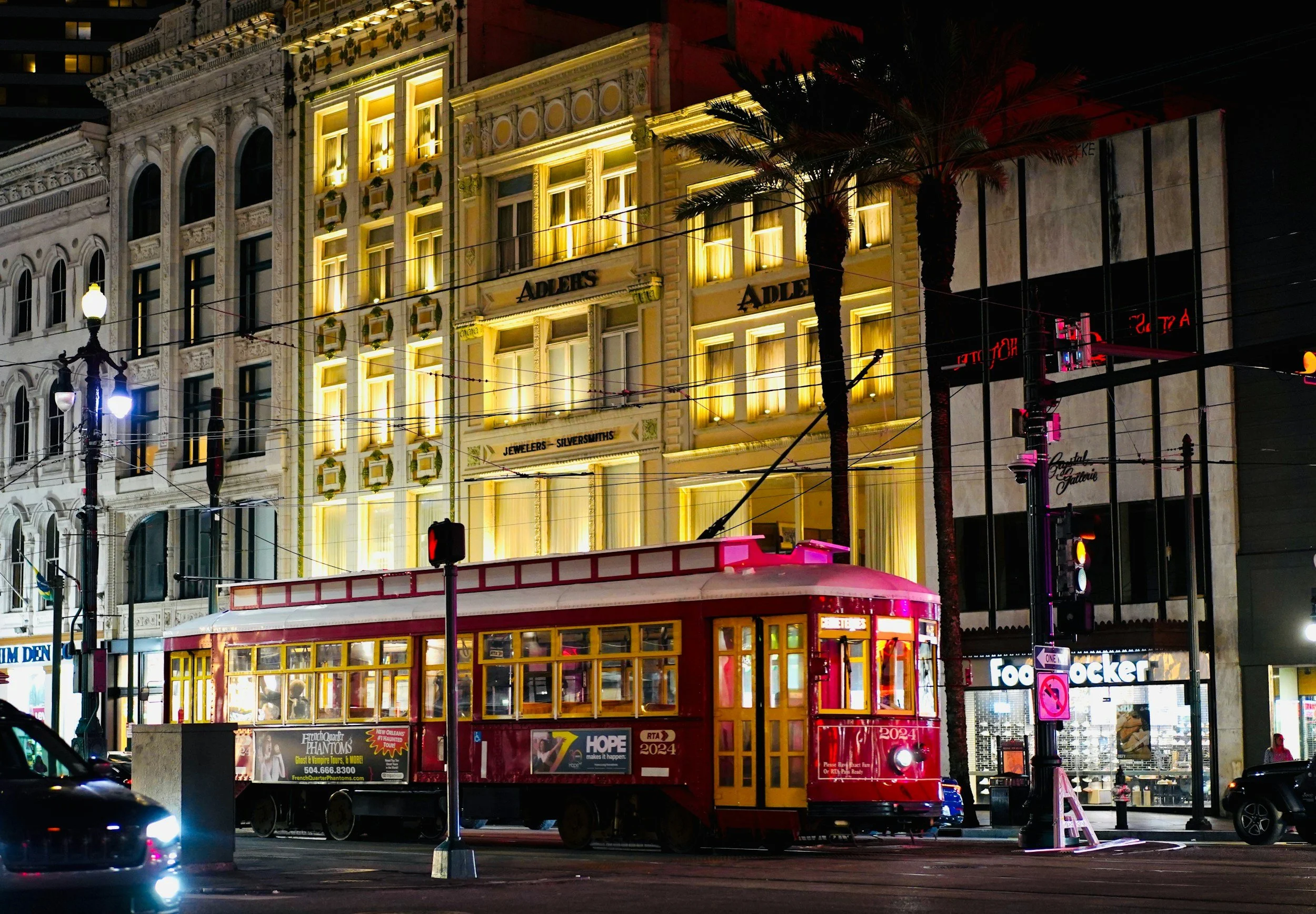 Red New Orleans streetcar traveling along Canal Street at night, passing historic buildings illuminated in warm golden lights with overhead streetcar wires and palm trees visible.
