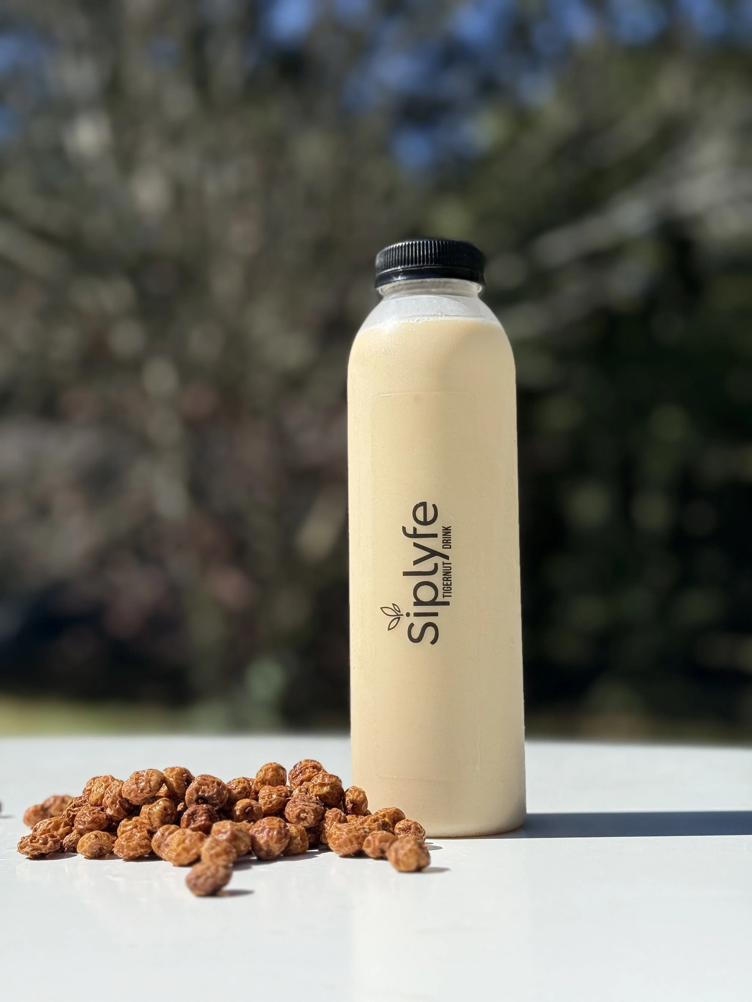 A bottle of Siplyfe tigernut drink with a small pile of dried tigernuts on a white surface outdoors, with blurred trees in the background.