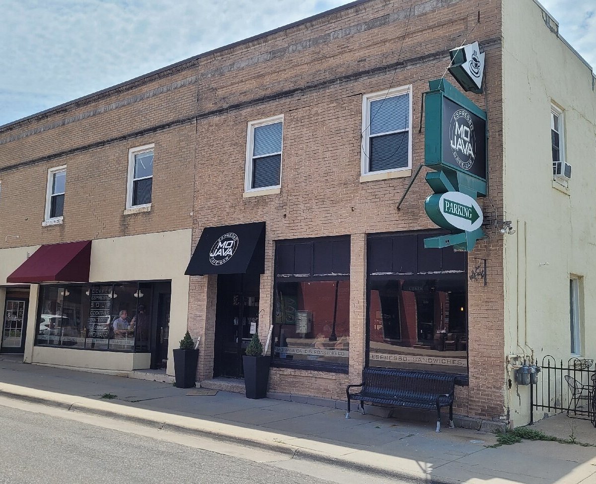 Brick building housing a Java coffee shop with a green and black sign, large windows, black awning, potted plants outside, and a parking sign mounted on the side.