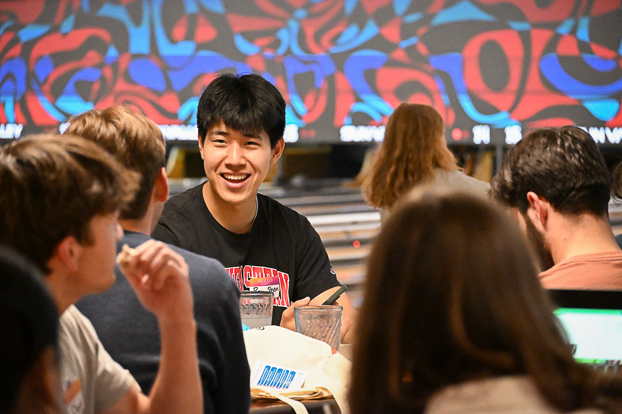 A group of people seated at a table in a social setting, with a young man smiling and engaging in conversation, and others around him, in front of a colorful abstract background.