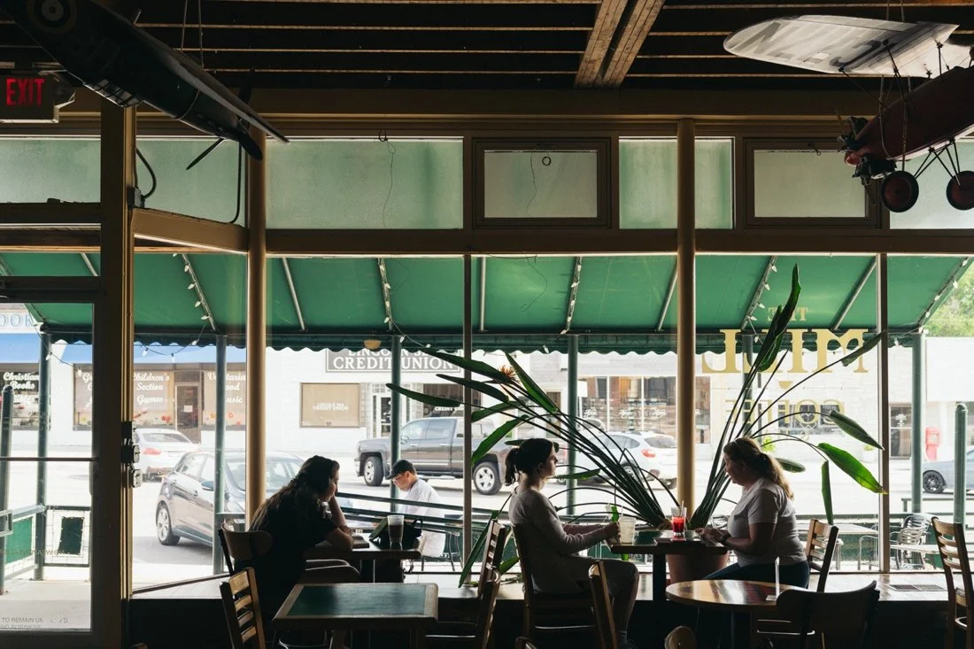 People are sitting at tables inside the Mill near the front large windows, with cars visible outside and green awnings overhead.