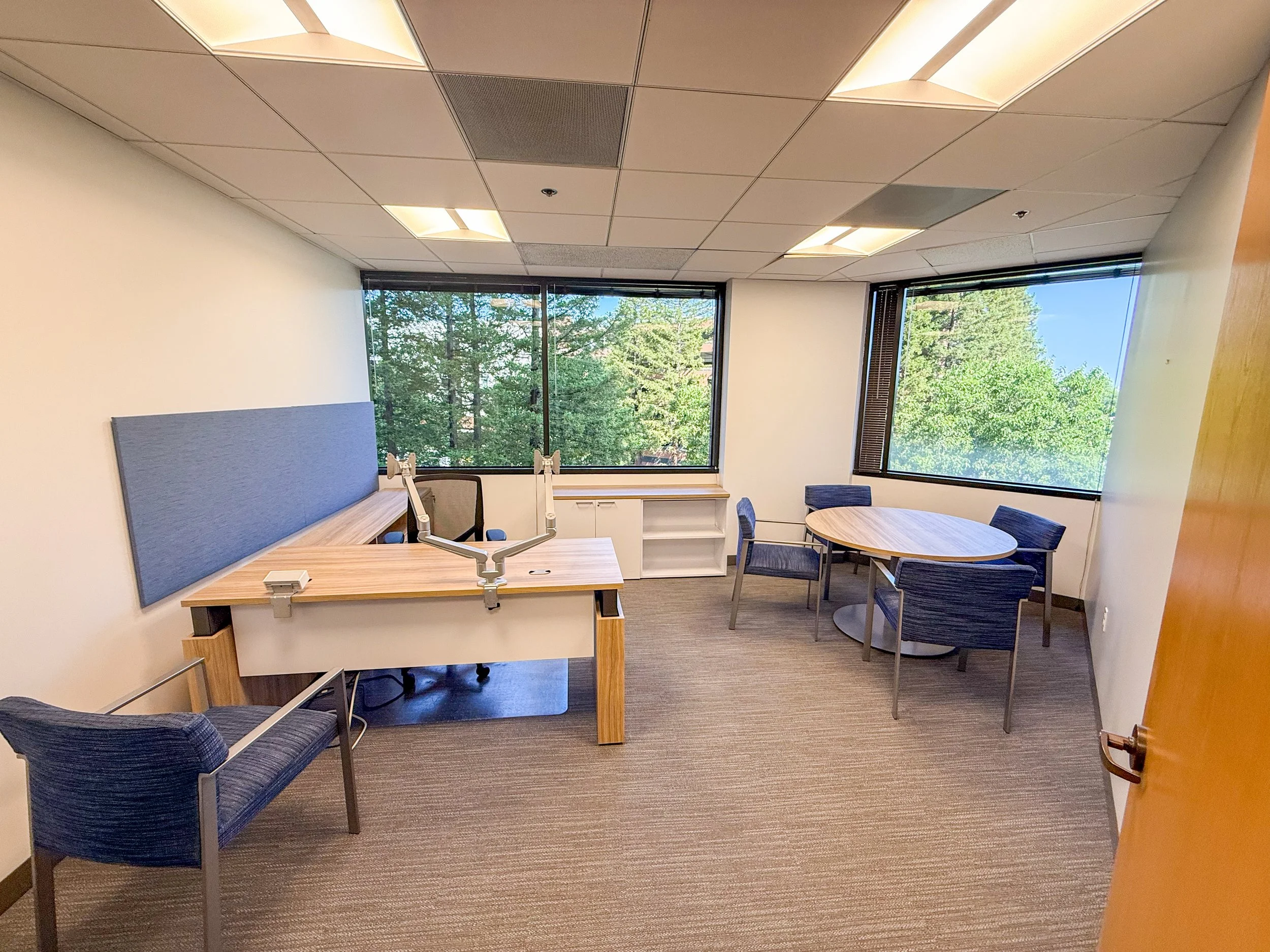 Empty office room with a desk and swivel chair, two wall-mounted whiteboards, a round table with four chairs, large windows showing trees outside, and a partially opened wooden door.