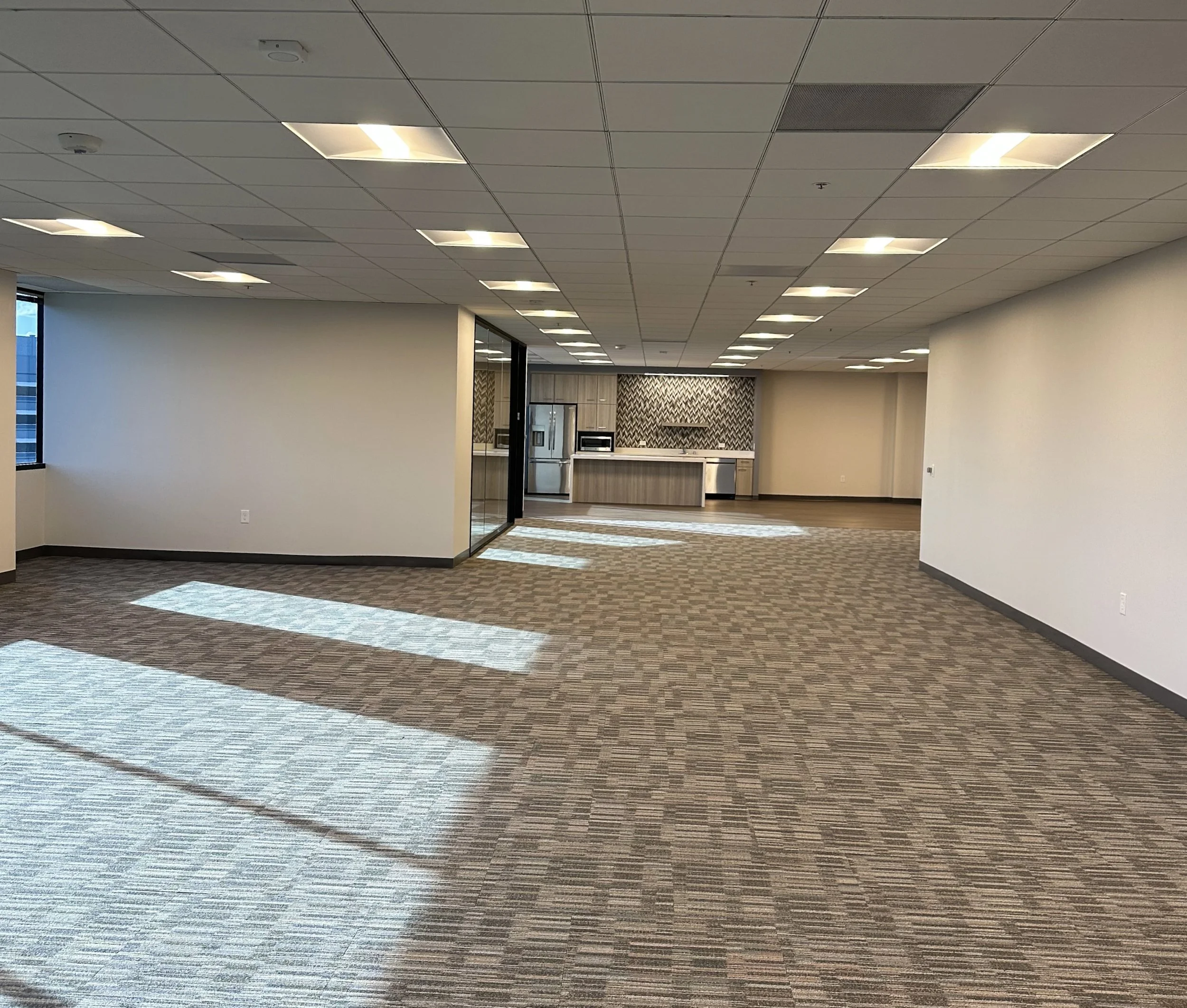 Empty office space with carpeting, white walls, ceiling lights, and a kitchenette with refrigerator, microwave, and patterned backsplash in the background.