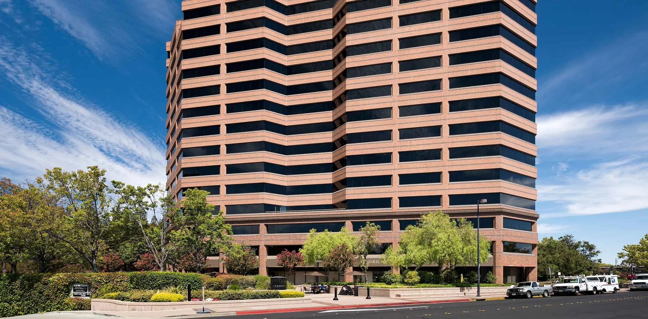 A tall, modern building with horizontal bands of windows and brick exterior, surrounded by trees and parked cars, under a partly cloudy blue sky.