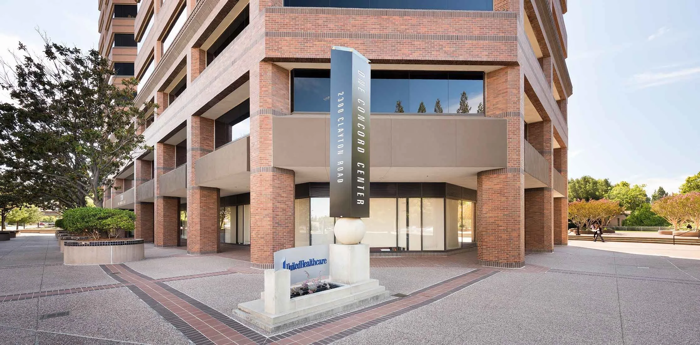 Exterior of a multi-story brick building with a vertical sign that reads 'Gervais Way, One Covington Road, 2300 Clayton Road.' A smaller sign at the ground level displays 'United Healthcare.' The area has trees and benches, with a clear sky.