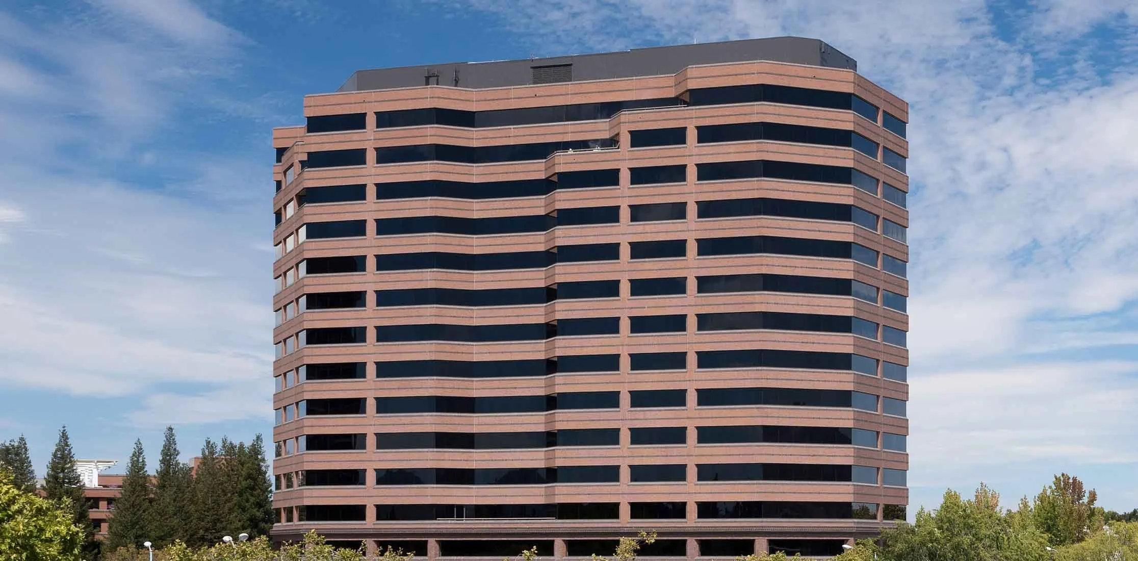 A tall modern office building with horizontal bands of dark tinted windows and pinkish-red brick exterior, surrounded by trees and a partly cloudy sky.