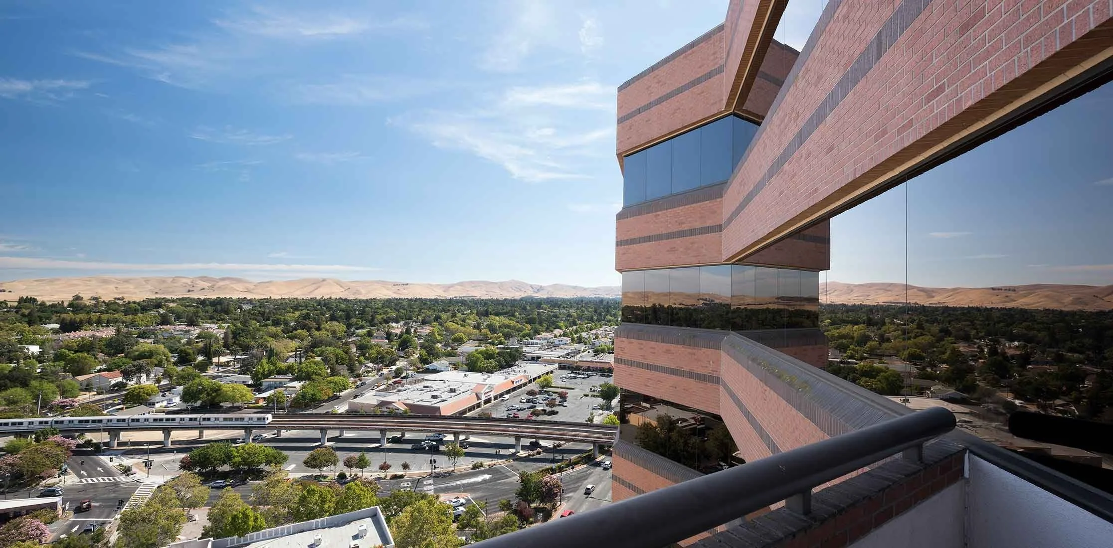 View from a balcony of a cityscape with trees, a highway with a train, and a brick building with reflective windows under a partly cloudy sky.
