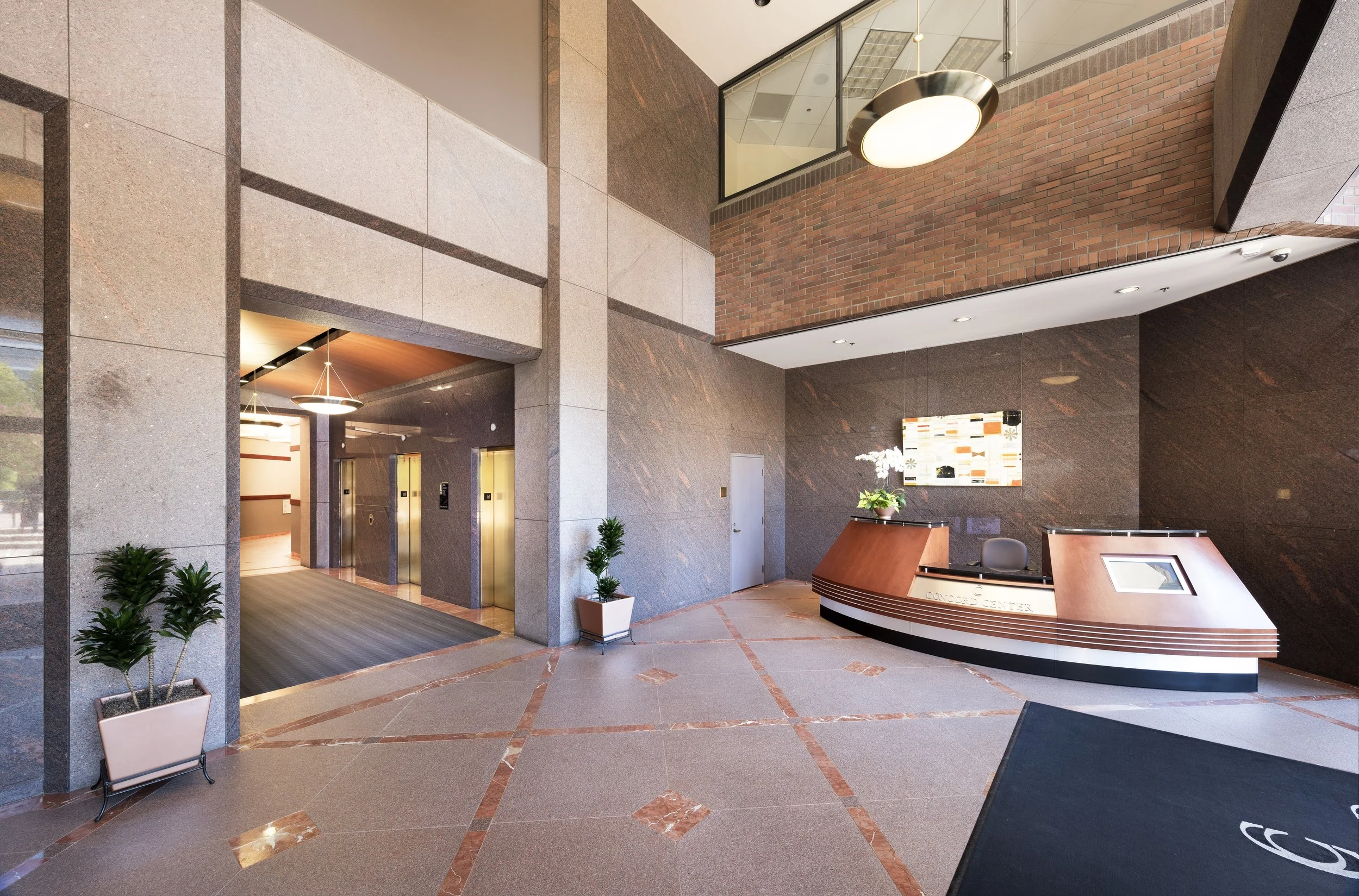 Lobby of a modern office building with a reception desk, elevator doors, potted plants, and artwork on the wall.