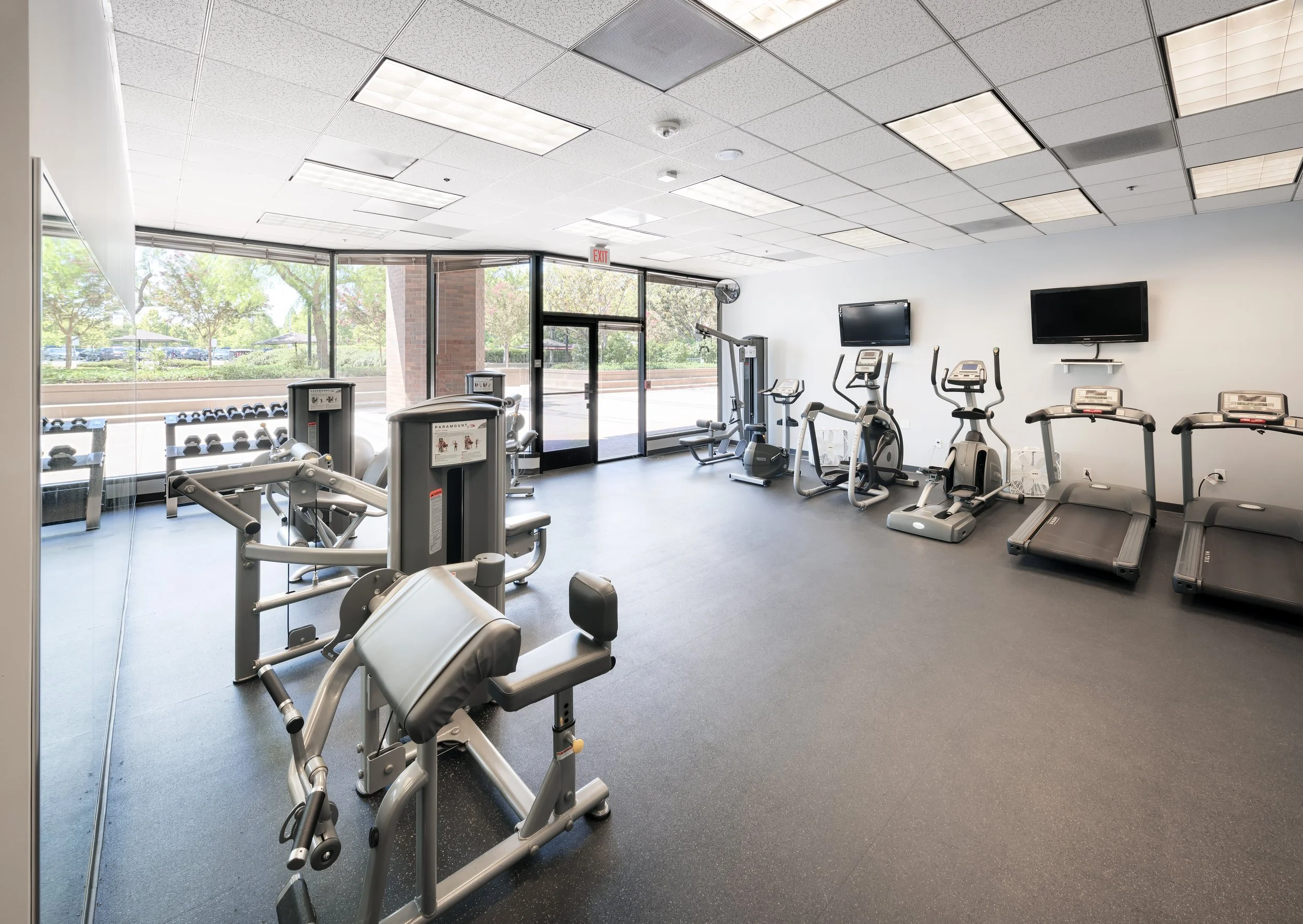 Empty fitness room with exercise machines, treadmills, stationary bikes, and weight equipment near large windows with outdoor view.