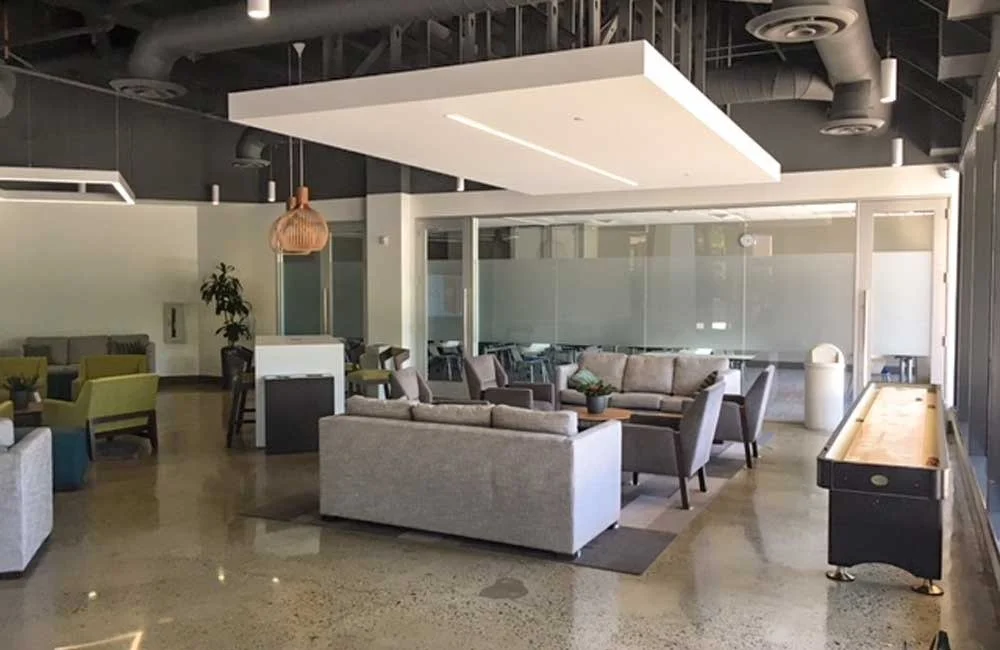 Modern lounge area with beige sofas, armchairs, a shuffleboard table, a large white ceiling panel, and large glass windows.