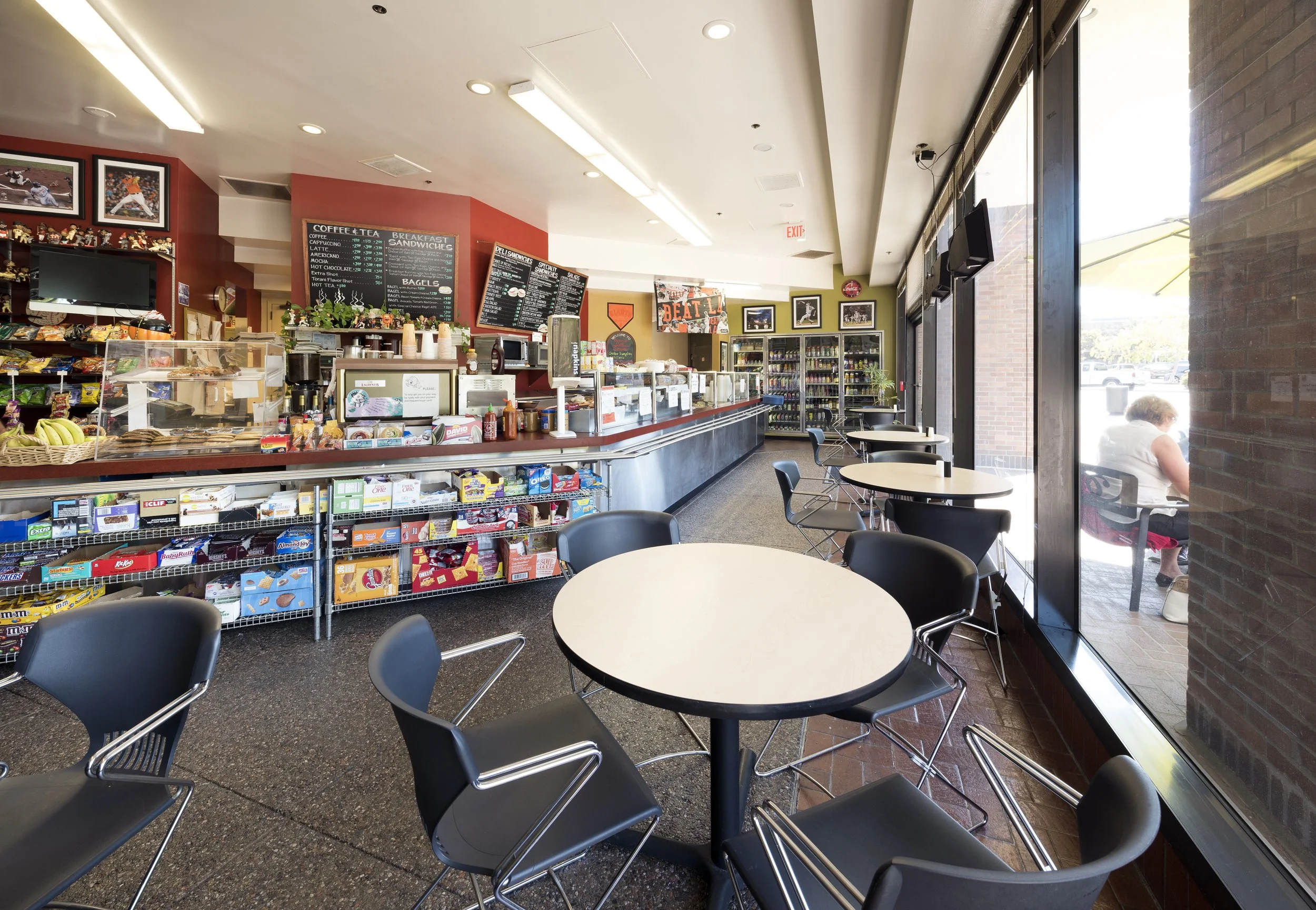 Interior of a coffee shop with round tables and black chairs near large windows, snack shelves near the counter, and a menu board behind the counter.