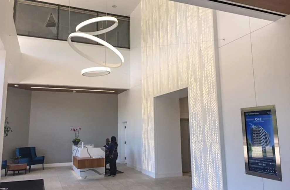Modern hotel lobby with white walls, a large illuminated decorative wall, a white reception desk with two people behind it, and a large circular hanging light fixture.