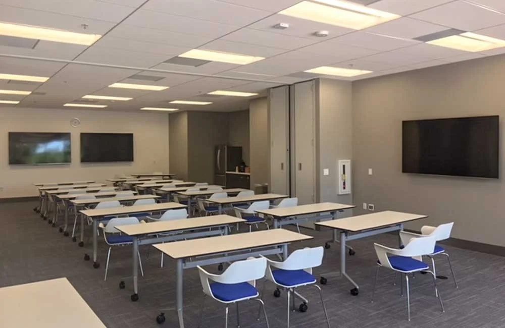 Empty classroom with rows of tables and chairs, large wall-mounted screens, and a kitchenette in the back.