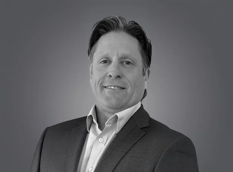 Black and white portrait of a smiling middle-aged man with dark, combed-back hair, wearing a dark suit jacket and a light collared shirt, against a plain background.