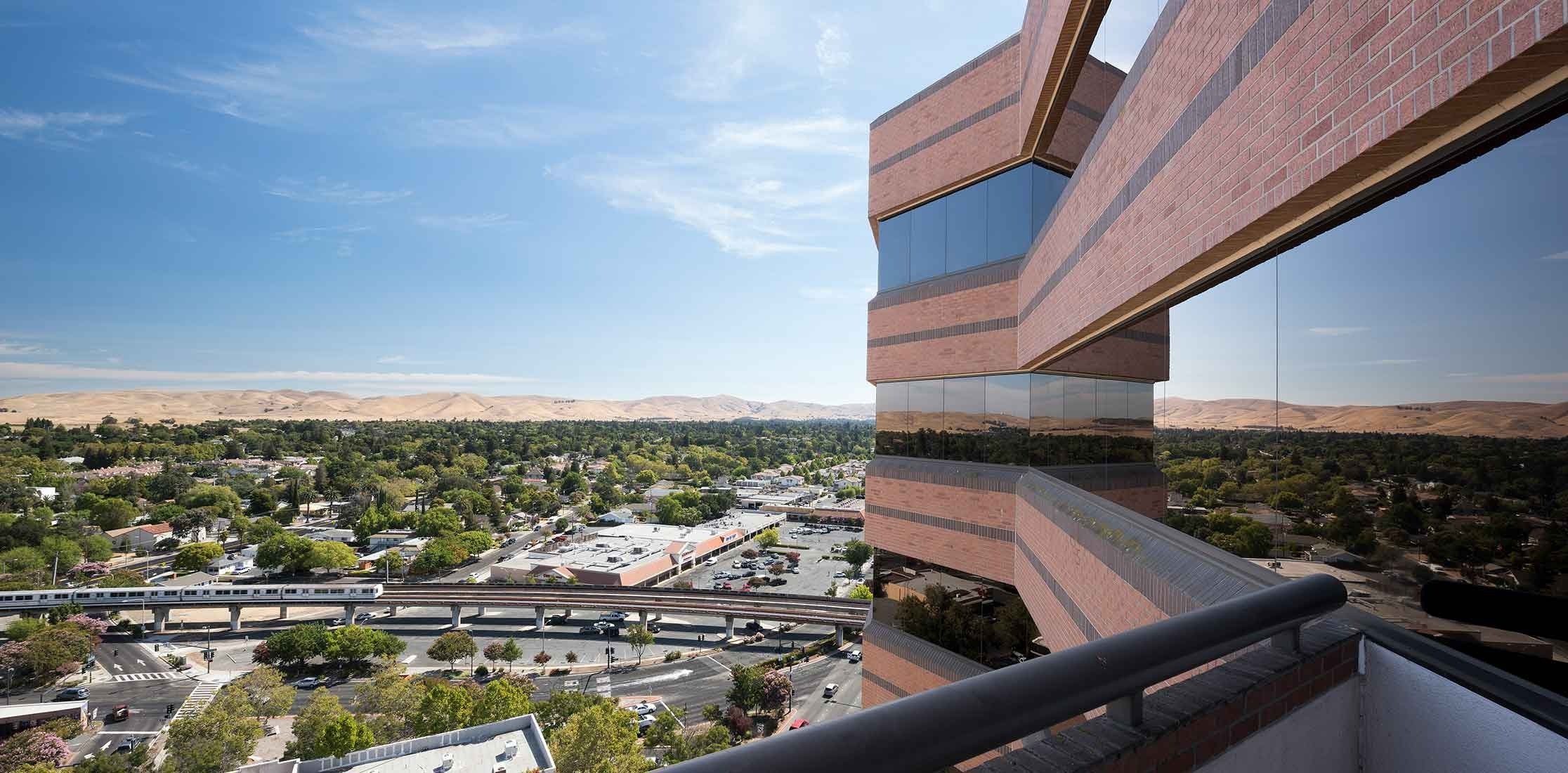 View from a high-rise building's balcony showing a cityscape with trees, a highway with a train, shopping centers, and distant hills under a clear blue sky.