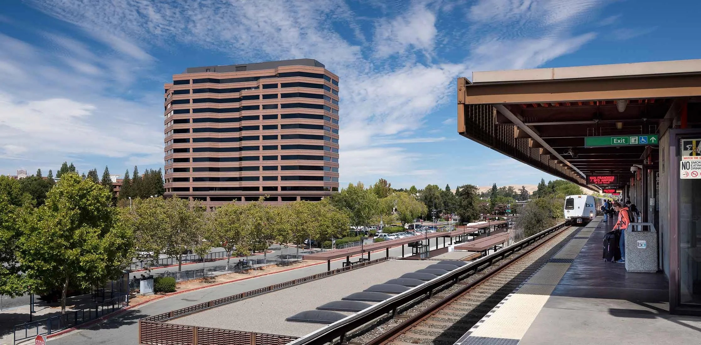 An elevated train station with a white train arriving, passengers waiting on the platform, a green exit sign, a cityscape with a large pinkish-brown building, trees, and a parking lot under a partly cloudy sky.