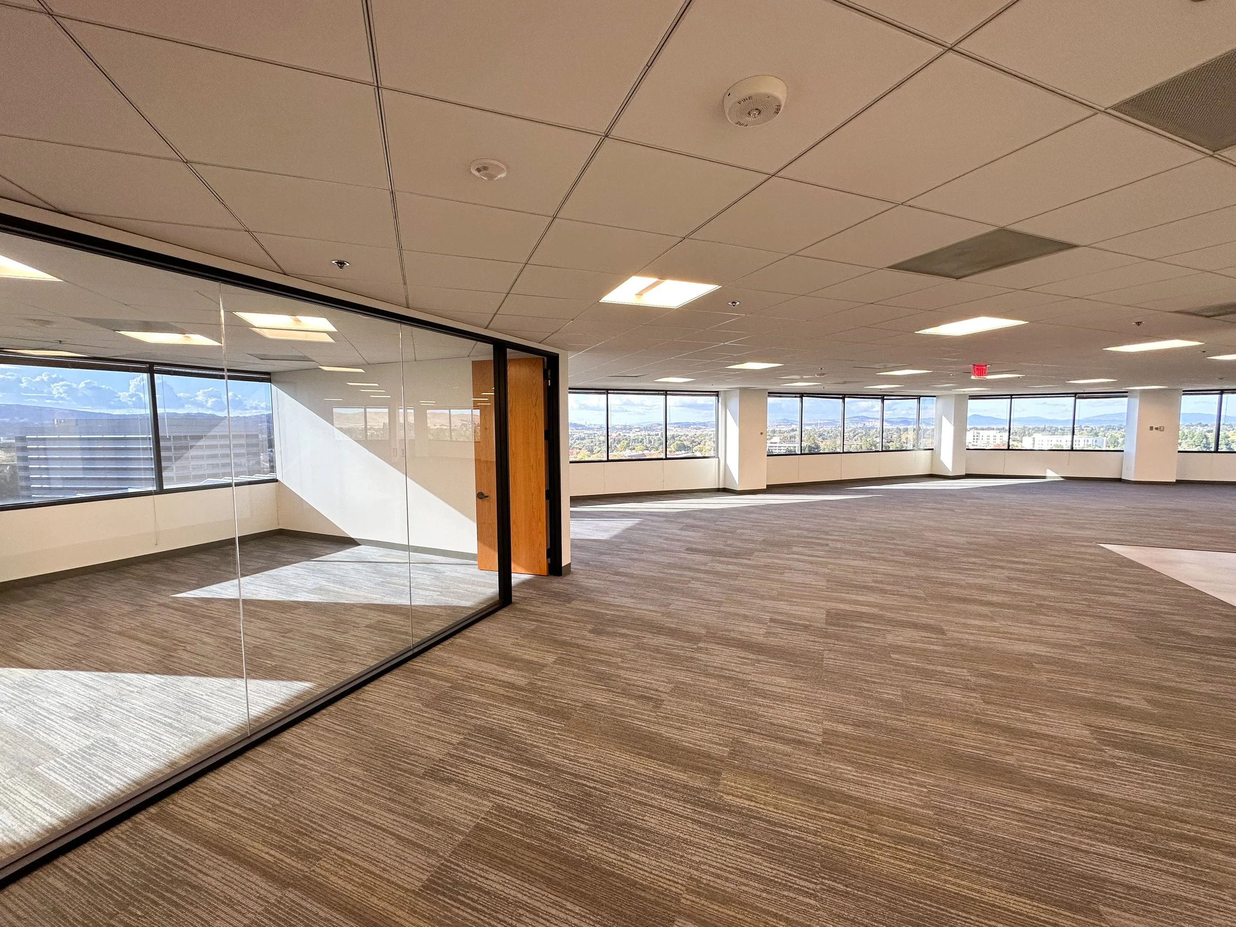 Empty office space with large windows, carpeted floor, and a glass walled conference room.