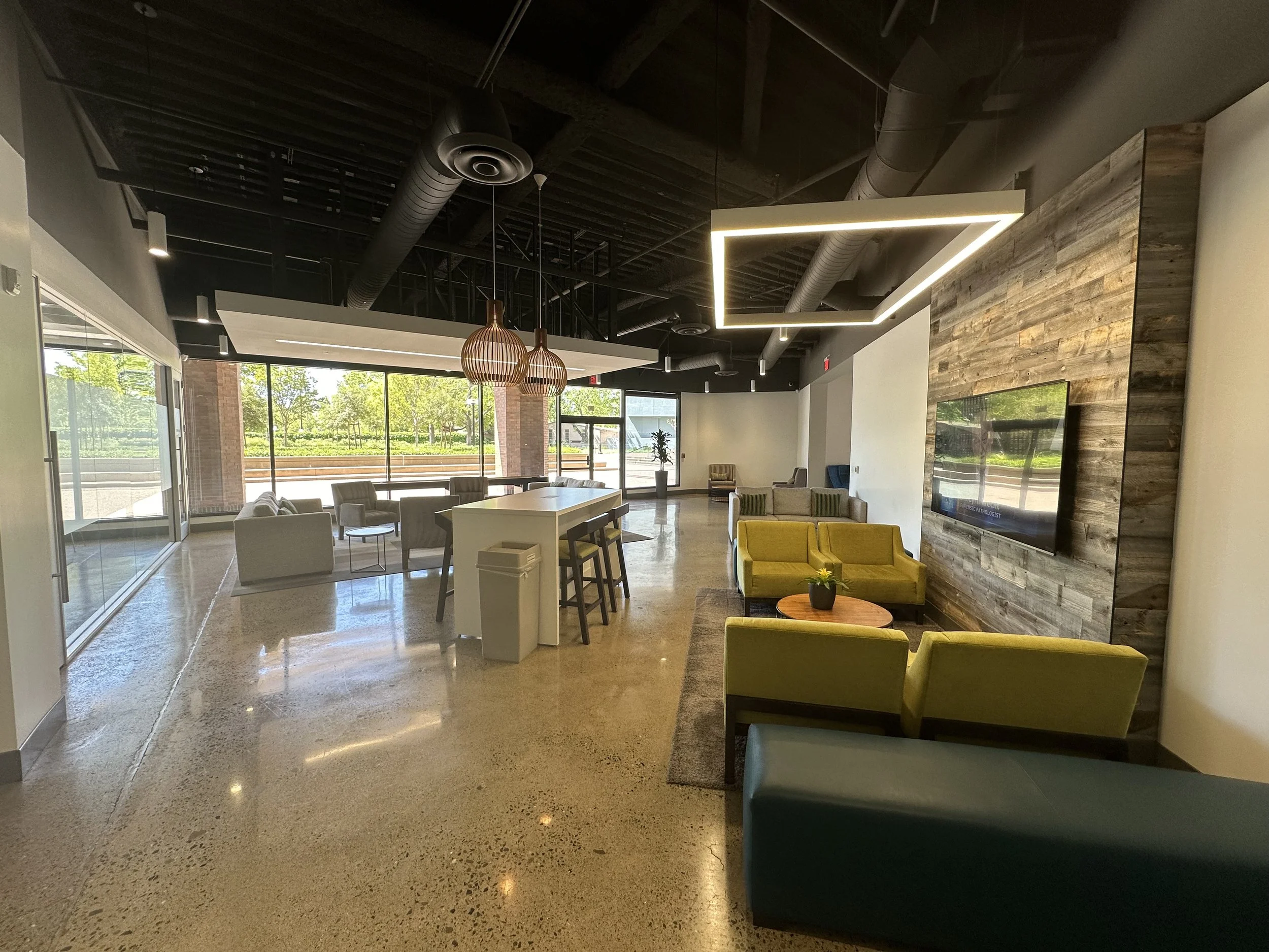 Modern lounge area with vibrant yellow and teal chairs, a large wall-mounted TV, and large windows showing trees outside.