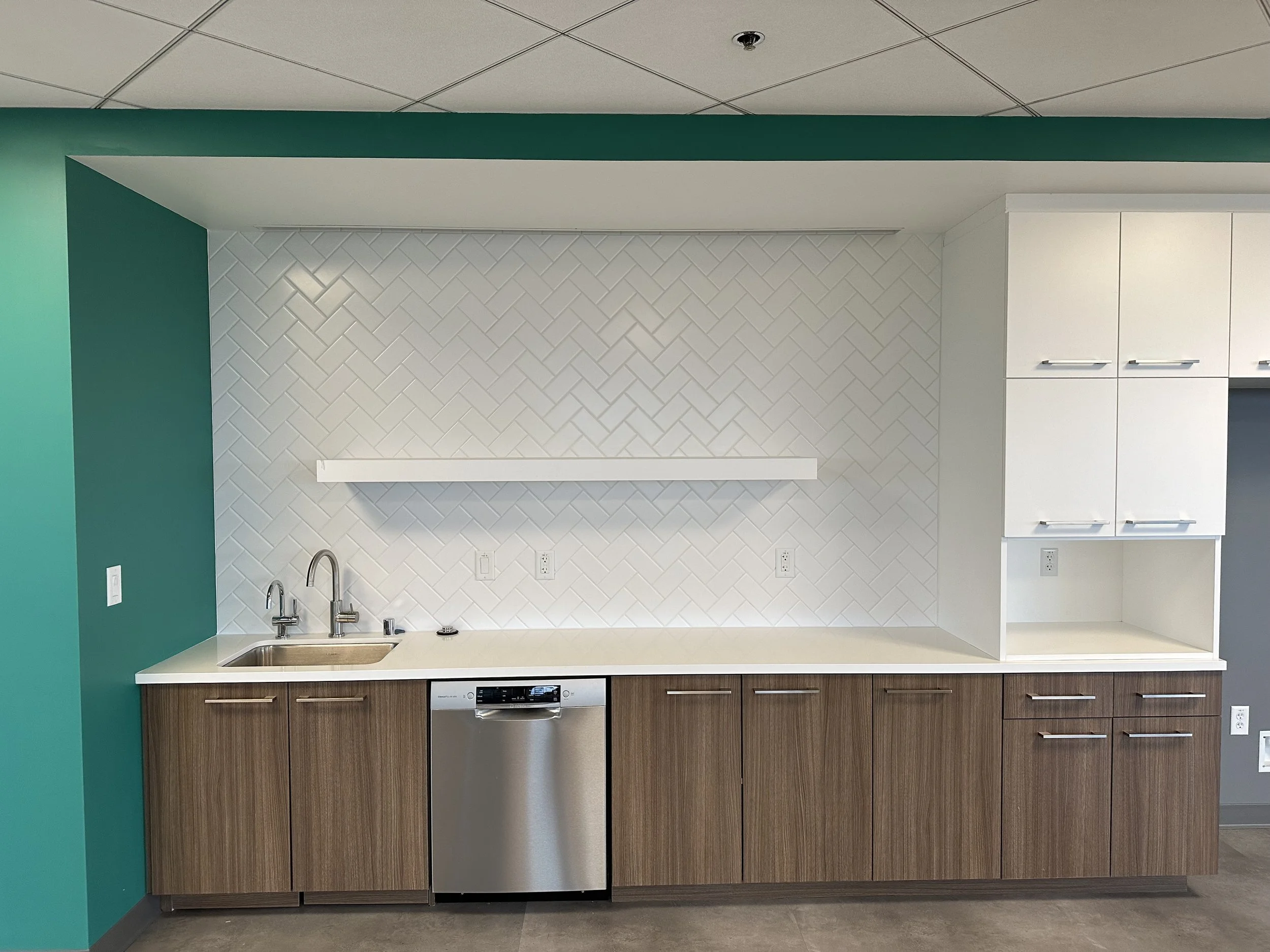 Modern kitchenette with white countertop, wood cabinetry, white textured tile backsplash, teal accent wall, stainless steel dishwasher, and white upper cabinets.