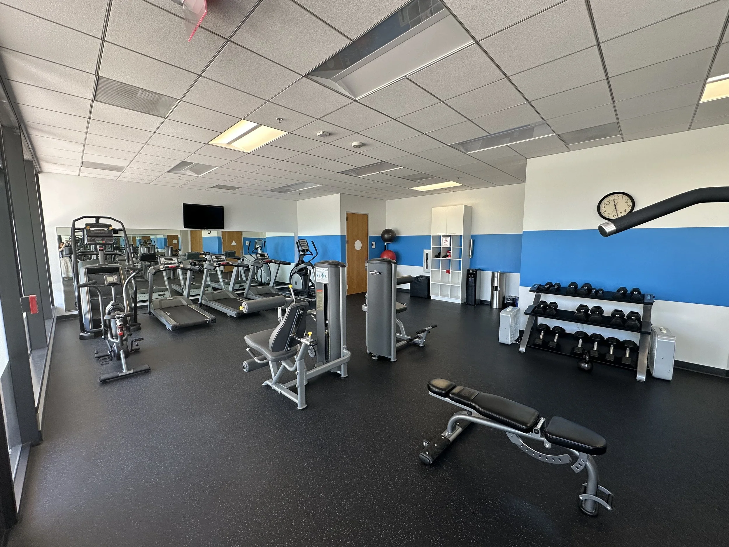 Empty workout room with various exercise machines, free weights, and a wall clock.