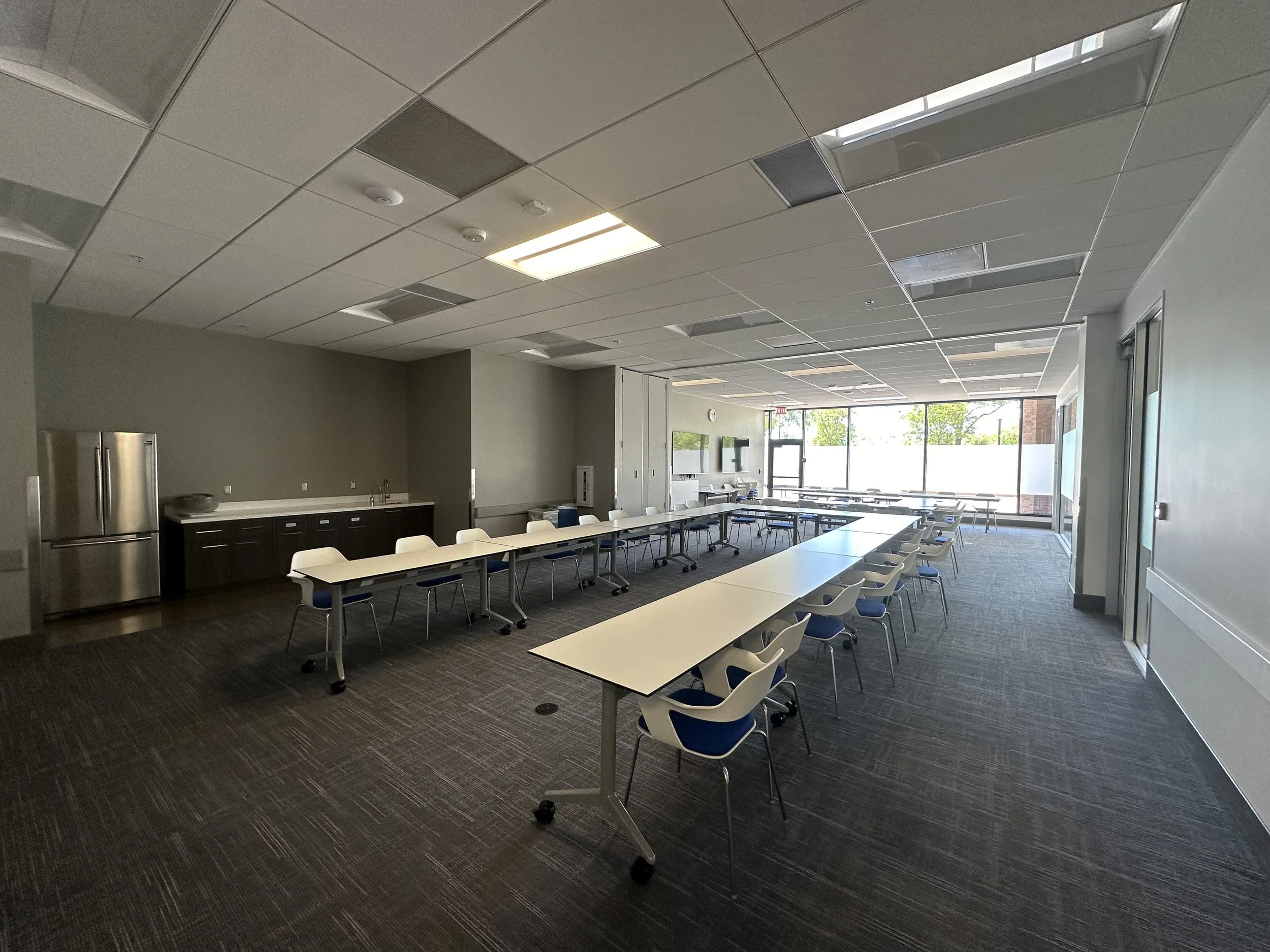 Empty conference room with white tables, white chairs with blue seats, large windows, and ceiling lights.