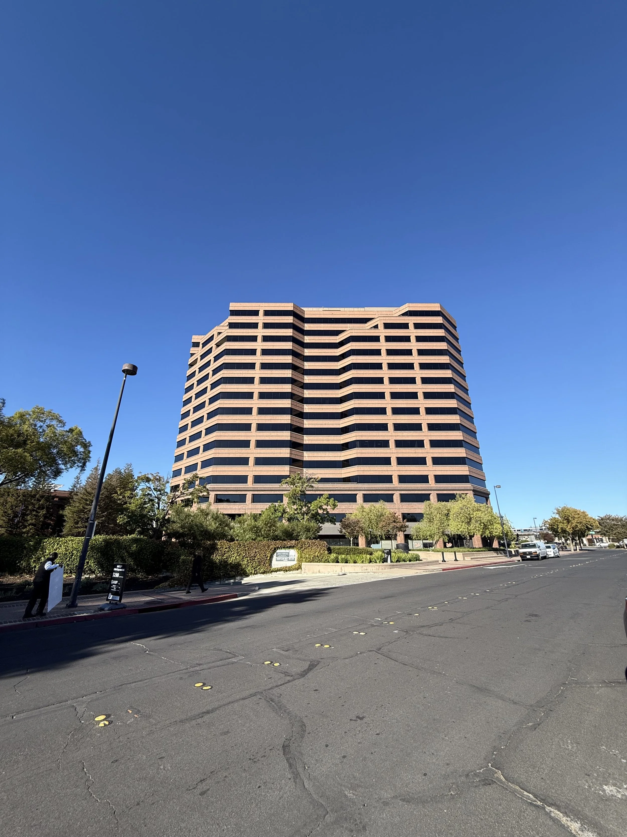 A large modern office building with a geometric design, surrounded by trees and a parking lot, under a clear blue sky.