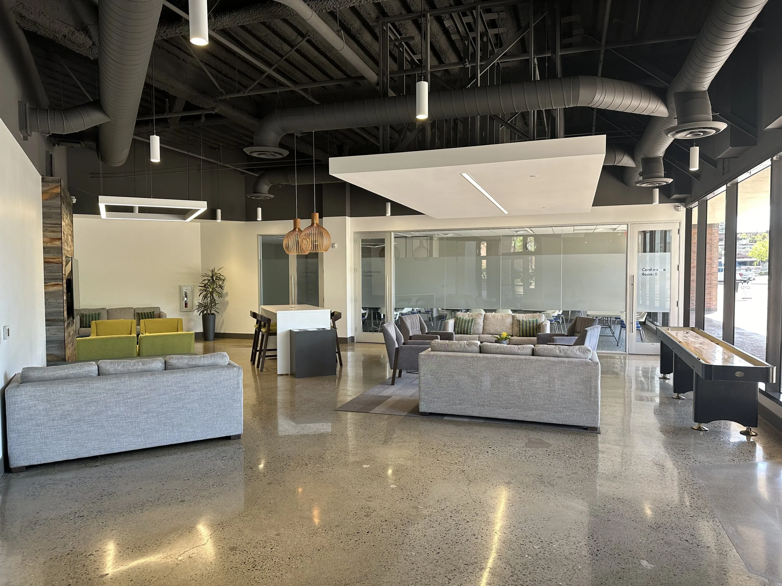 Modern lobby with couches, chairs, and large windows, featuring exposed ceiling ducts and contemporary lighting.