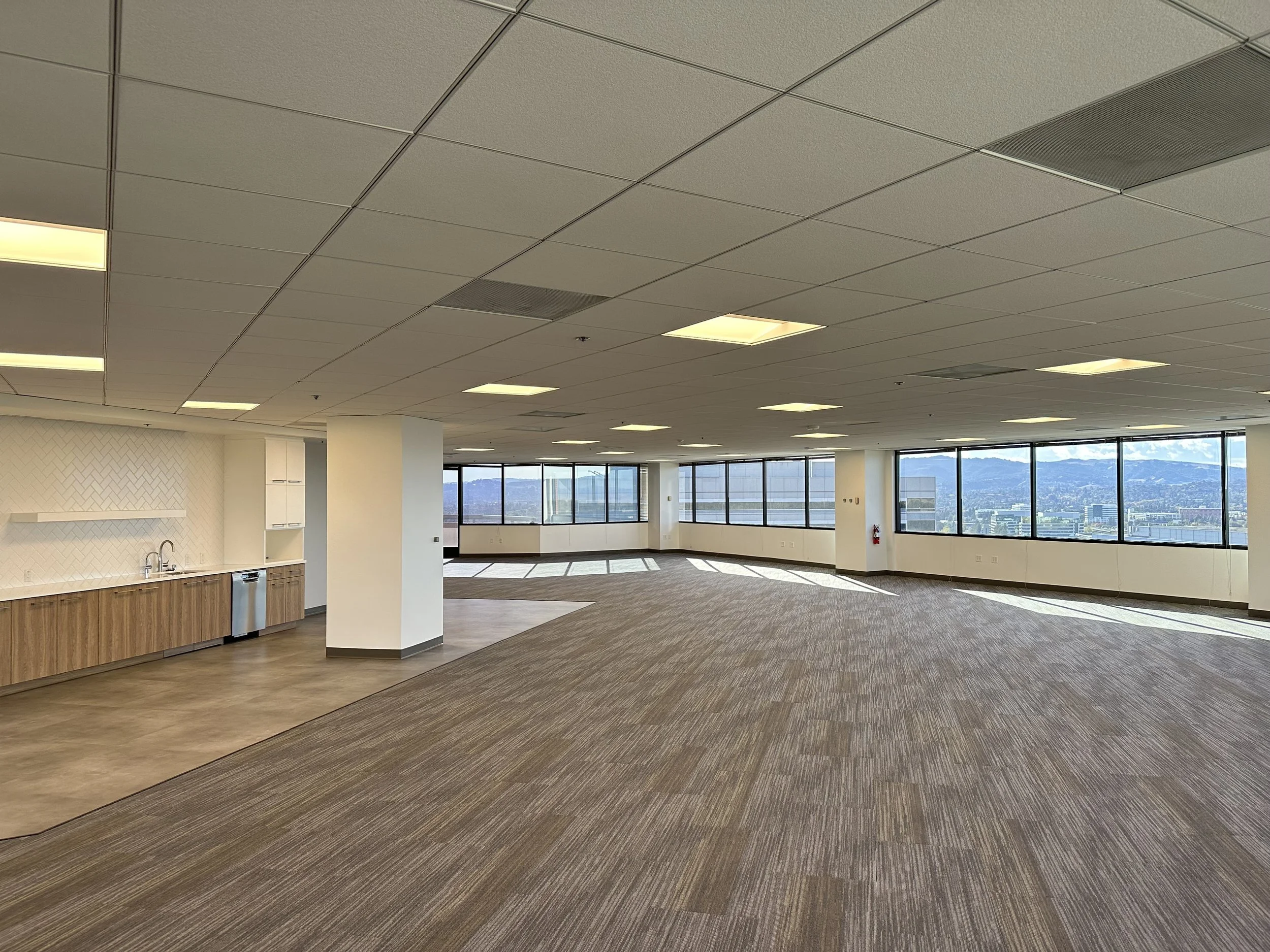 Spacious empty office space with large windows offering city views, a kitchenette area with cabinets and a sink, and a ceiling with grid panels and lighting.