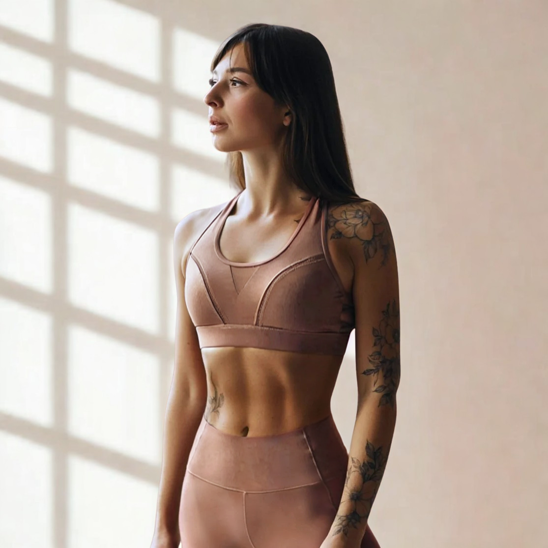 Woman with tattoos wearing a sports bra and leggings standing indoors near a window with sunlight casting shadows.