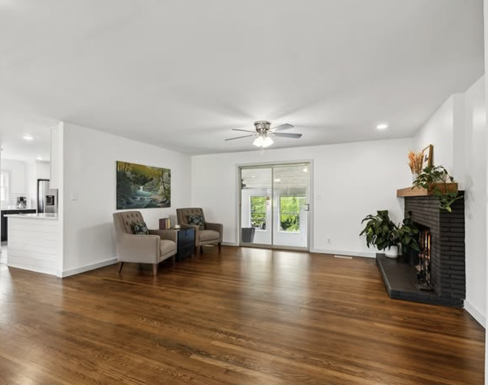 A spacious living room with white walls, a wooden floor, a ceiling fan, and a fireplace decorated with plants. There are two beige armchairs and a painting on the wall. Double sliding glass doors lead to an outdoor area.