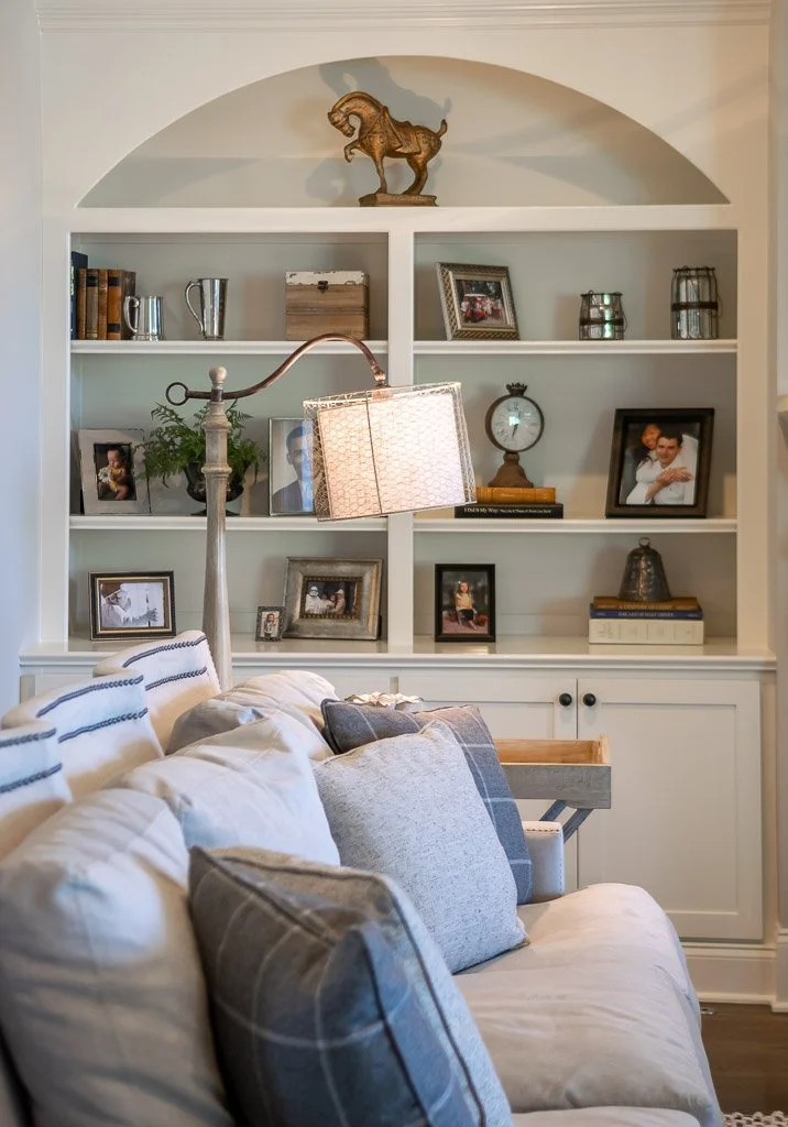 A living room with a white built-in shelf filled with family photos, books, and decorative items. There is a couch with various throw pillows in front of the shelf, and a floor lamp with a rectangular shade positioned beside the couch in the foreground.