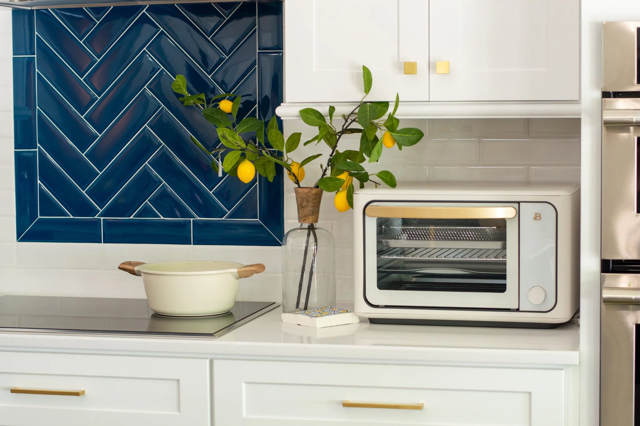 A kitchen countertop with a white oven, a leafy lemon branch in a glass vase, a beige ceramic dish with wooden handles, and white cabinetry with gold handles. There is a blue tiled backsplash with a herringbone pattern.