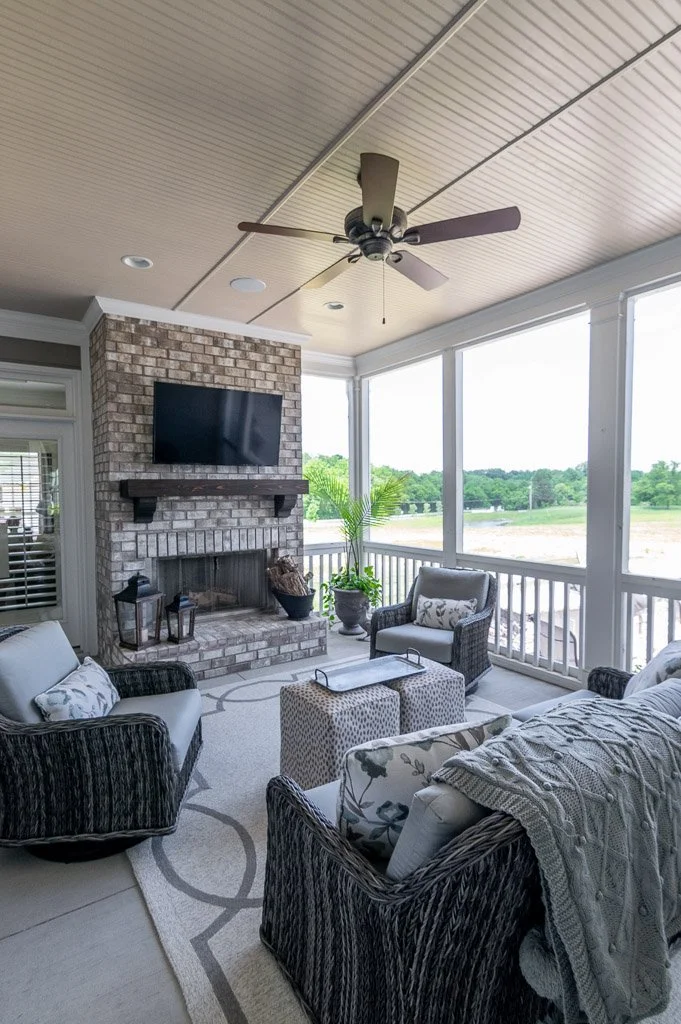 Cozy screened-in porch with wicker furniture, a brick fireplace, a wall-mounted TV, and large windows overlooking a green yard.