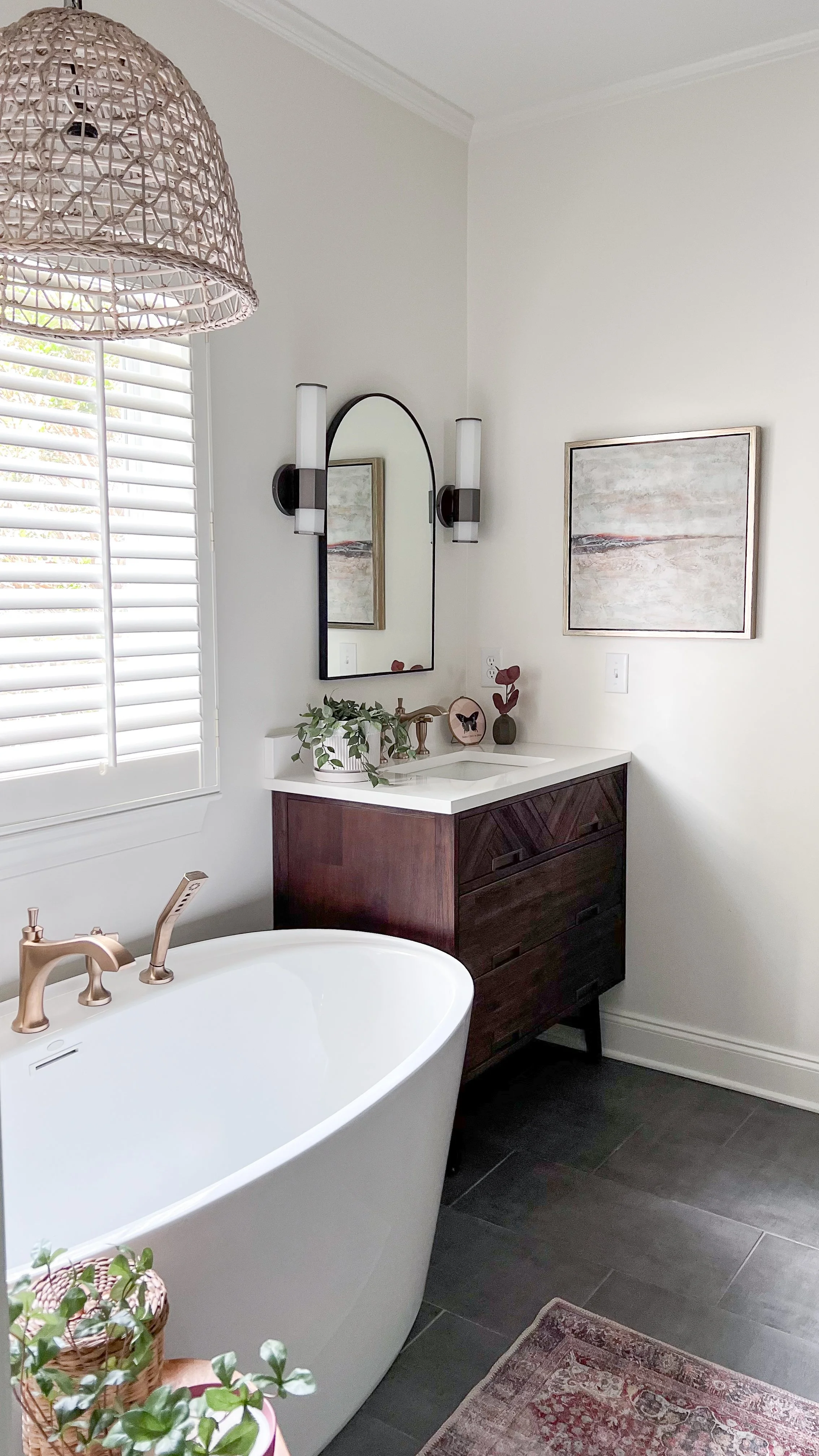 A modern bathroom with a white bathtub, a dark wood vanity with a white countertop, and a decorative mirror with wall sconces. There are also framed artwork, potted plants, and a window with white shutters.