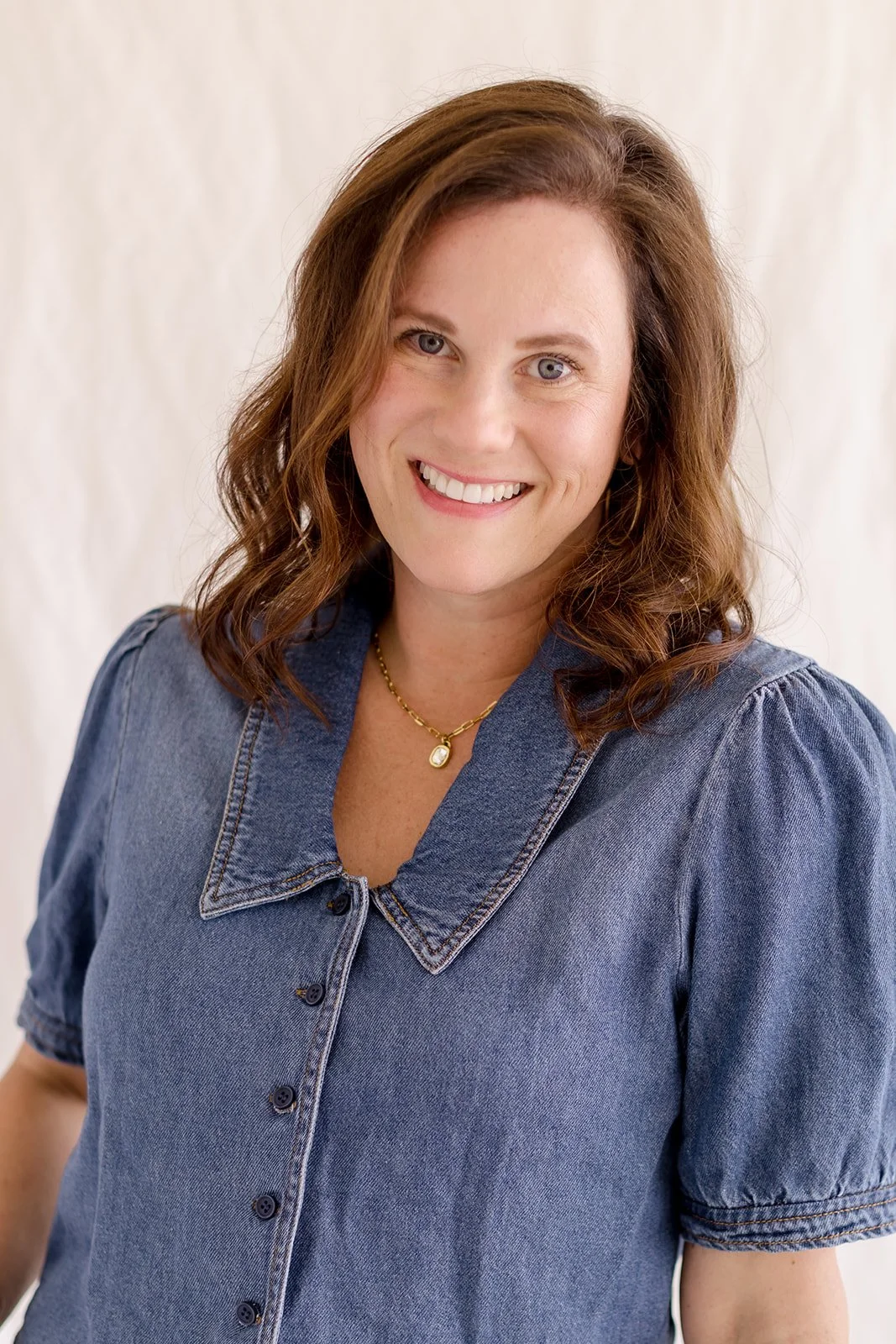 A woman with shoulder-length curly brown hair, fair skin, and blue eyes, smiling at the camera, wearing a denim shirt with puffed sleeves and a gold necklace with a pendant.