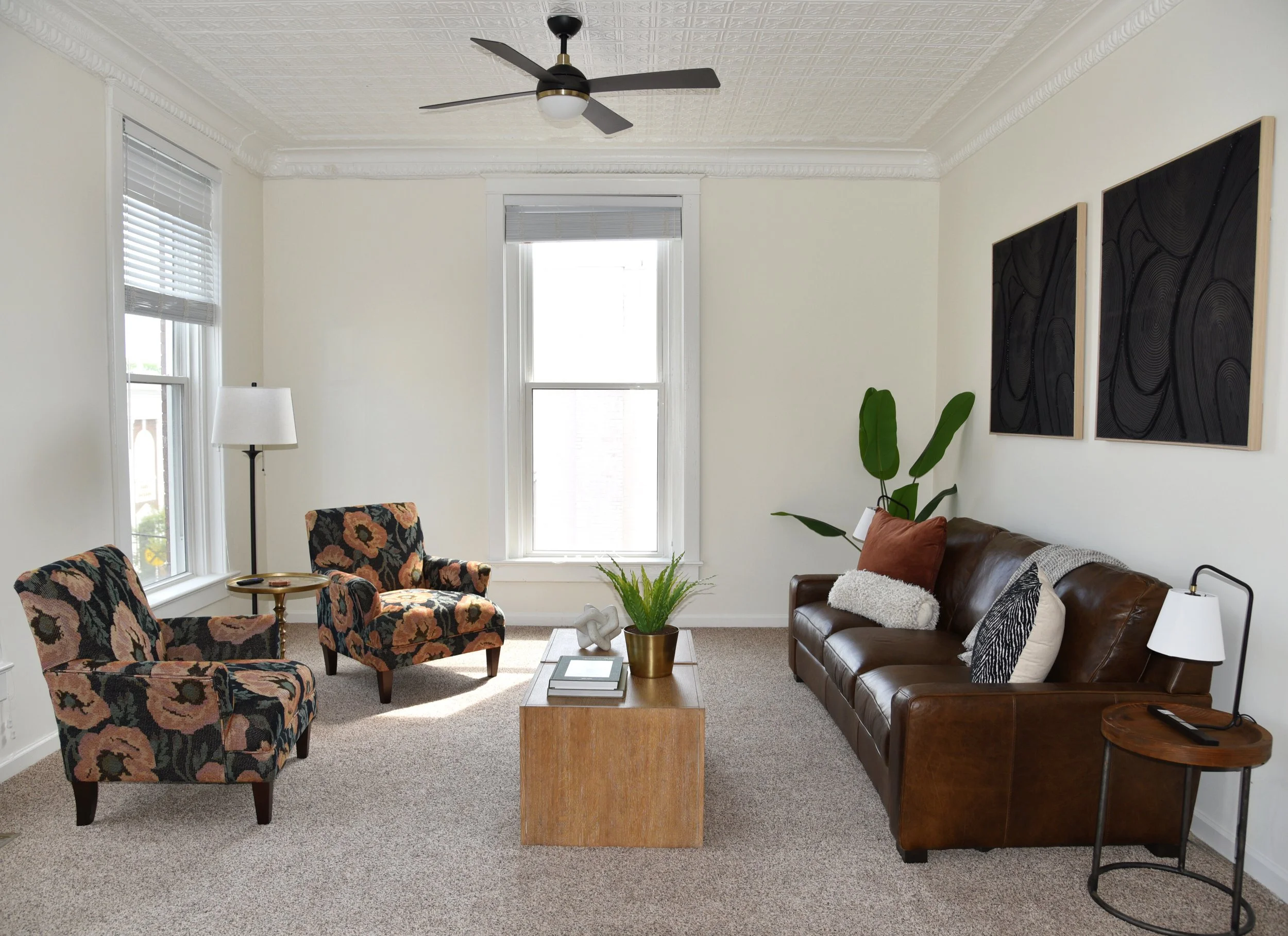 Living room with two patterned armchairs, a leather sofa, a wooden coffee table, a floor lamp, and wall art, featuring windows and a ceiling fan.