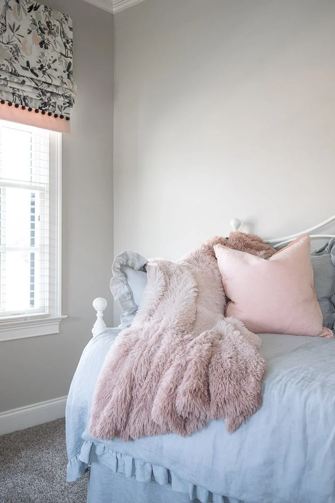 A cozy bedroom corner with a white bed, pink and gray pillows, a fluffy pink throw blanket, and a window with floral-patterned curtains.