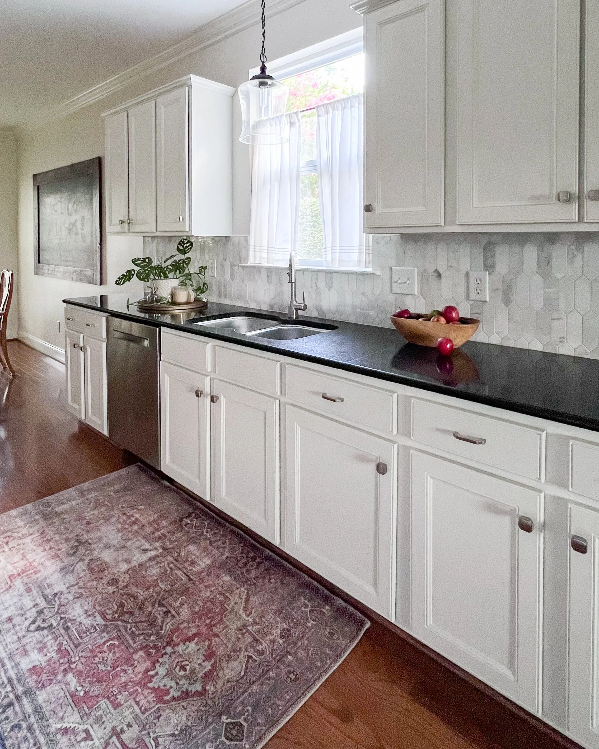 Kitchen with white cabinets, black countertop, window with white curtains, potted plant, wooden bowl of fruit, stainless steel dishwasher, and a decorative rug.