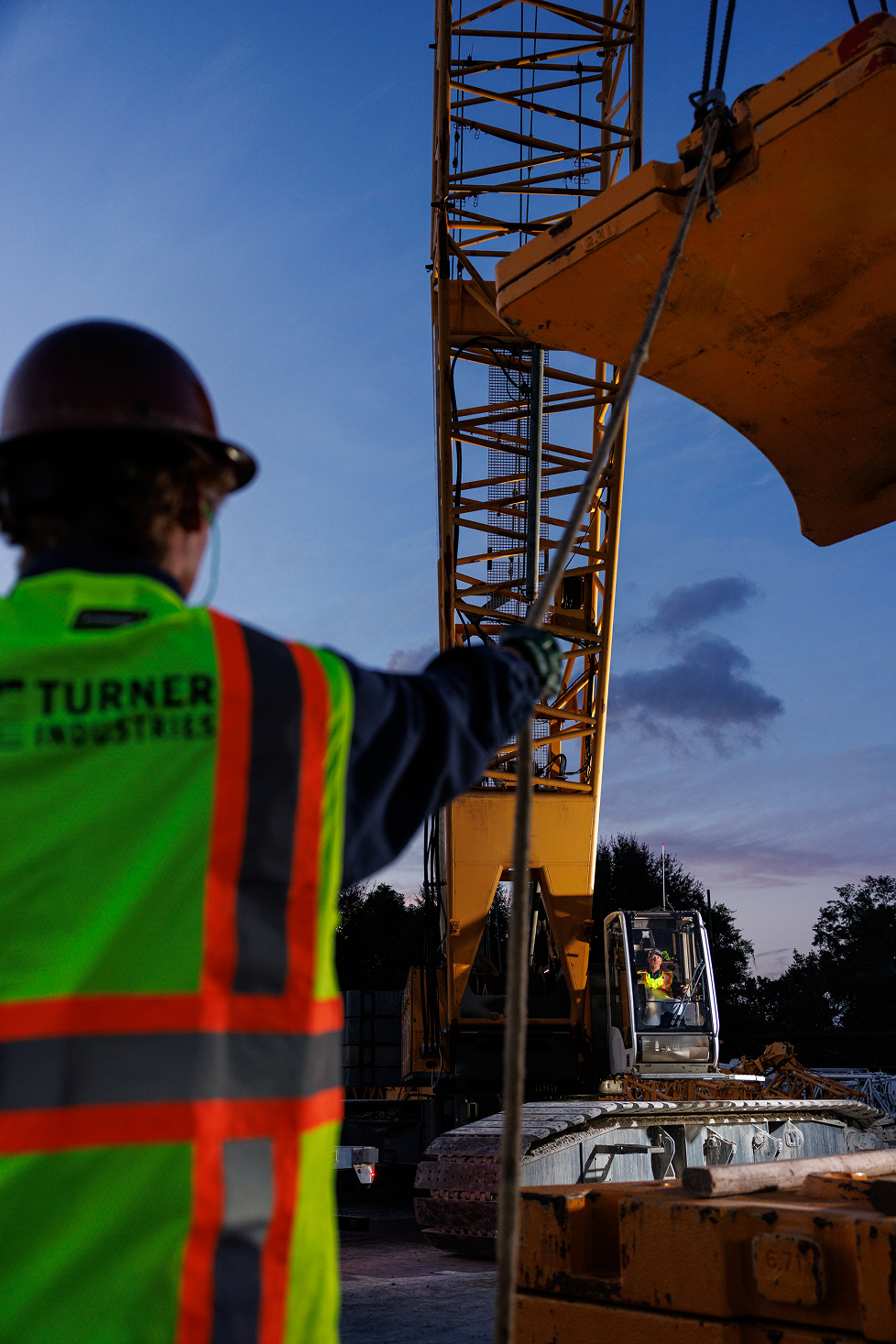 Construction worker in safety vest and helmet operating a crane during dusk.