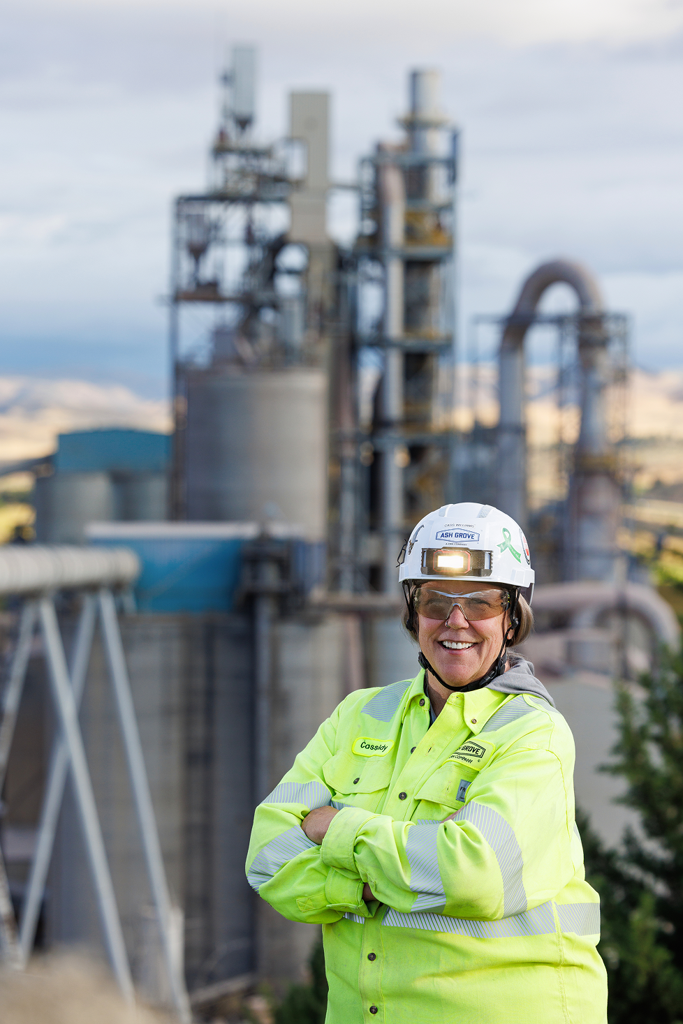 Smiling woman in safety gear standing in front of an industrial plant, with arms crossed.